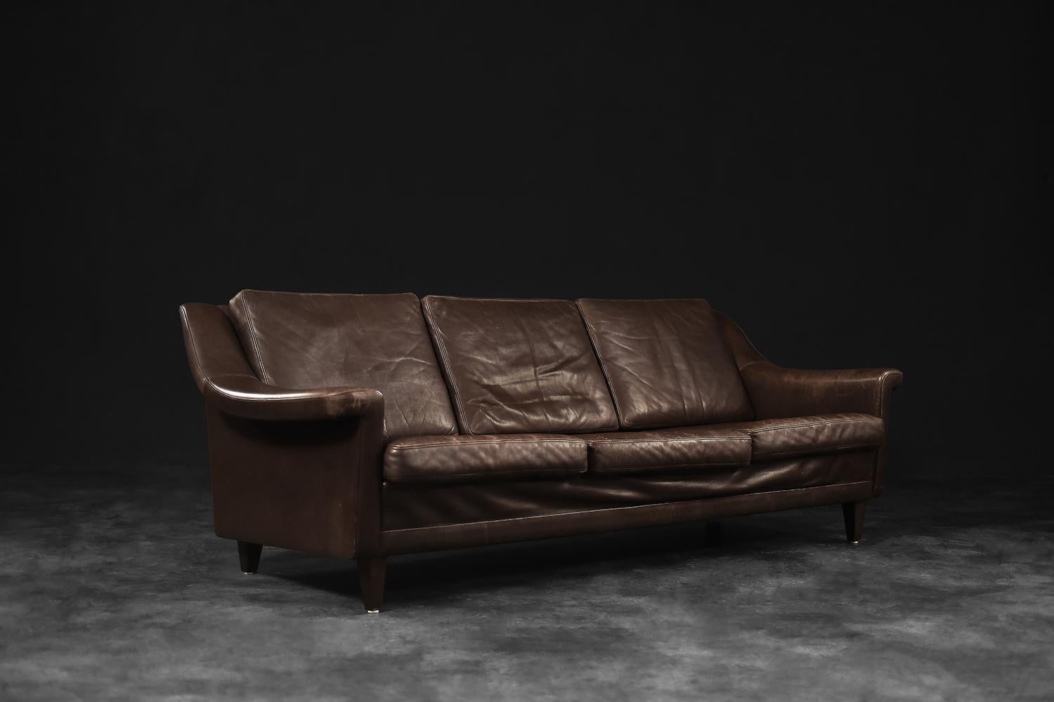This wide three-seater sofa was made in Denmark in the 1970s. The modernist frame with six pillows is upholstered in chestnut colored natural leather. The sofa has subtly rounded edges and armrests. The wide and comfortable seat makes the comfort of