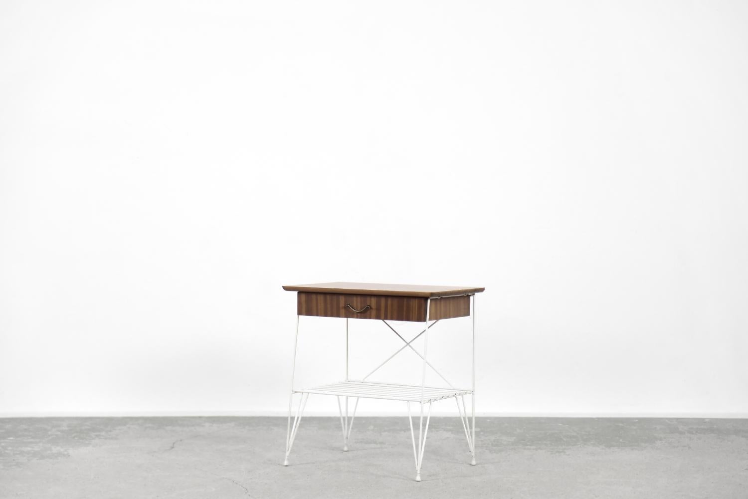 This modernist bedside table was made in Sweden during the 1960s. The top with the drawer is finished in walnut wood in a warm shade of brown. The metal legs, connected to the lower shelf, are in white, which makes them look good despite the passage