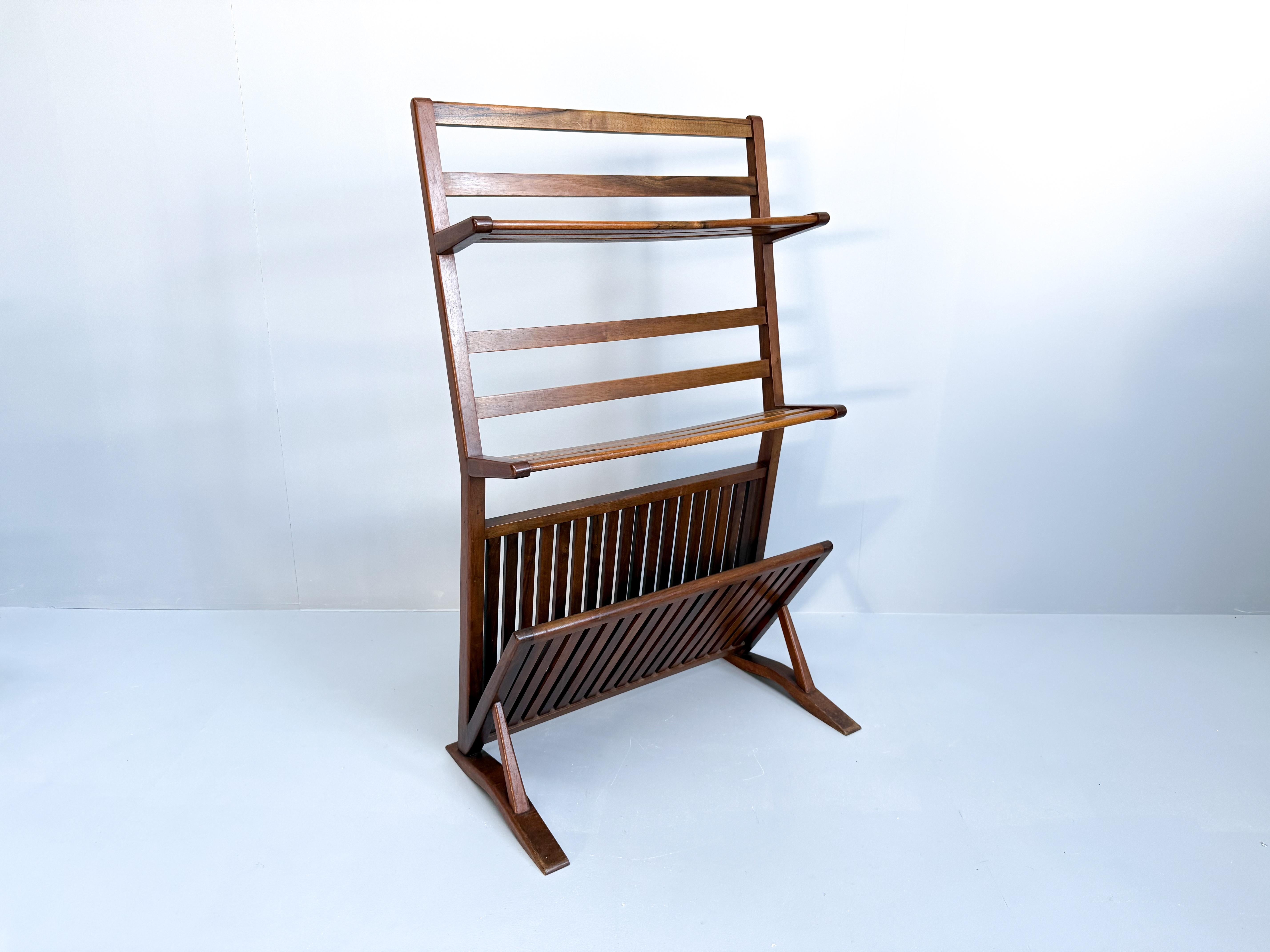 This mid-century modern bookshelf and magazine rack is made from walnut wood, dating back to the 1960s. The piece has a sleek and minimalistic design, featuring a slanted frame with horizontal wooden bars for holding books, and a lower compartment