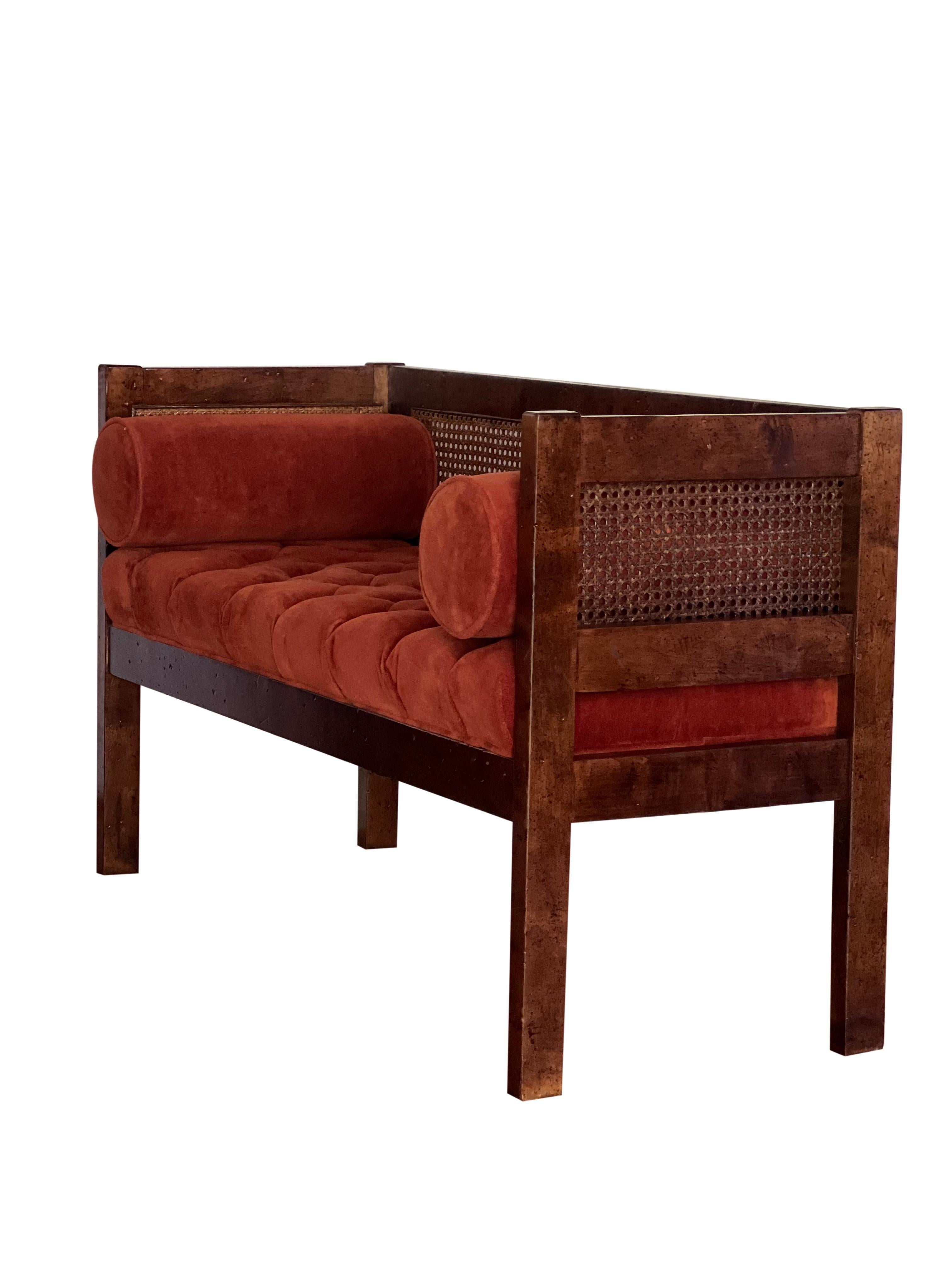 Elegant vintage Hollywood Regency cane parlor bench. 

The bench features button tufted velvet upholstery in persimmon with two matching bolsters. The seat cushion is attached to the piece. The cane back and sides are in good condition, no holes