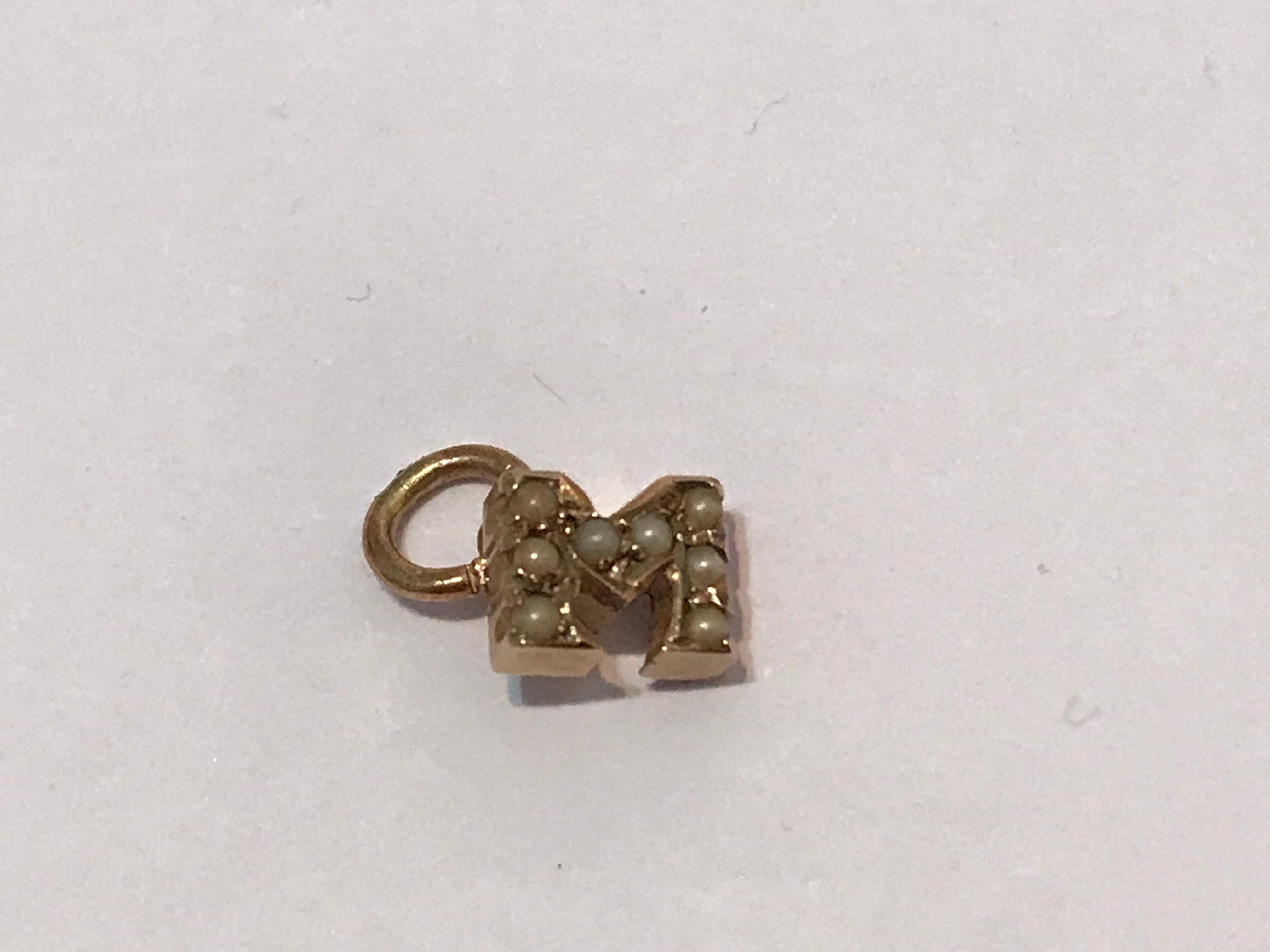 The tiny 14k rose gold initial M set with eight seed pearls attached to a gold suspension ring.  

Circa 1900

1/8 in. (.3 cm) high; 3/16 in. (.5 cm) wide; 1/16 in. (.2 cm) high. Chain not included. 
