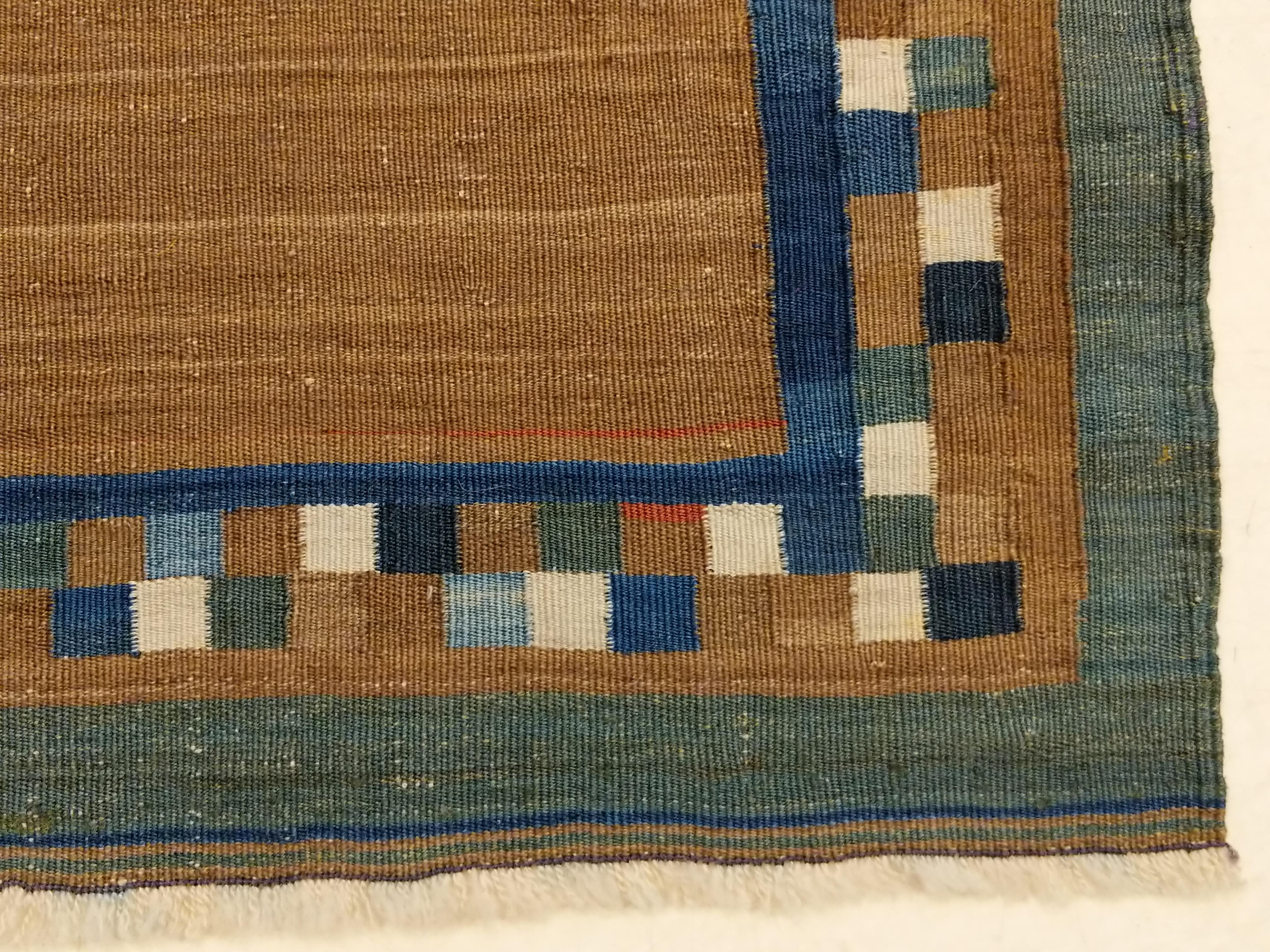A diminutive masterpiece of flat-woven tribal art, distinguished by a dazzling chequerboard border framing an open field composed of natural camel wool. The apparently random arrangement of polychrome squares is reminiscent of the most refined