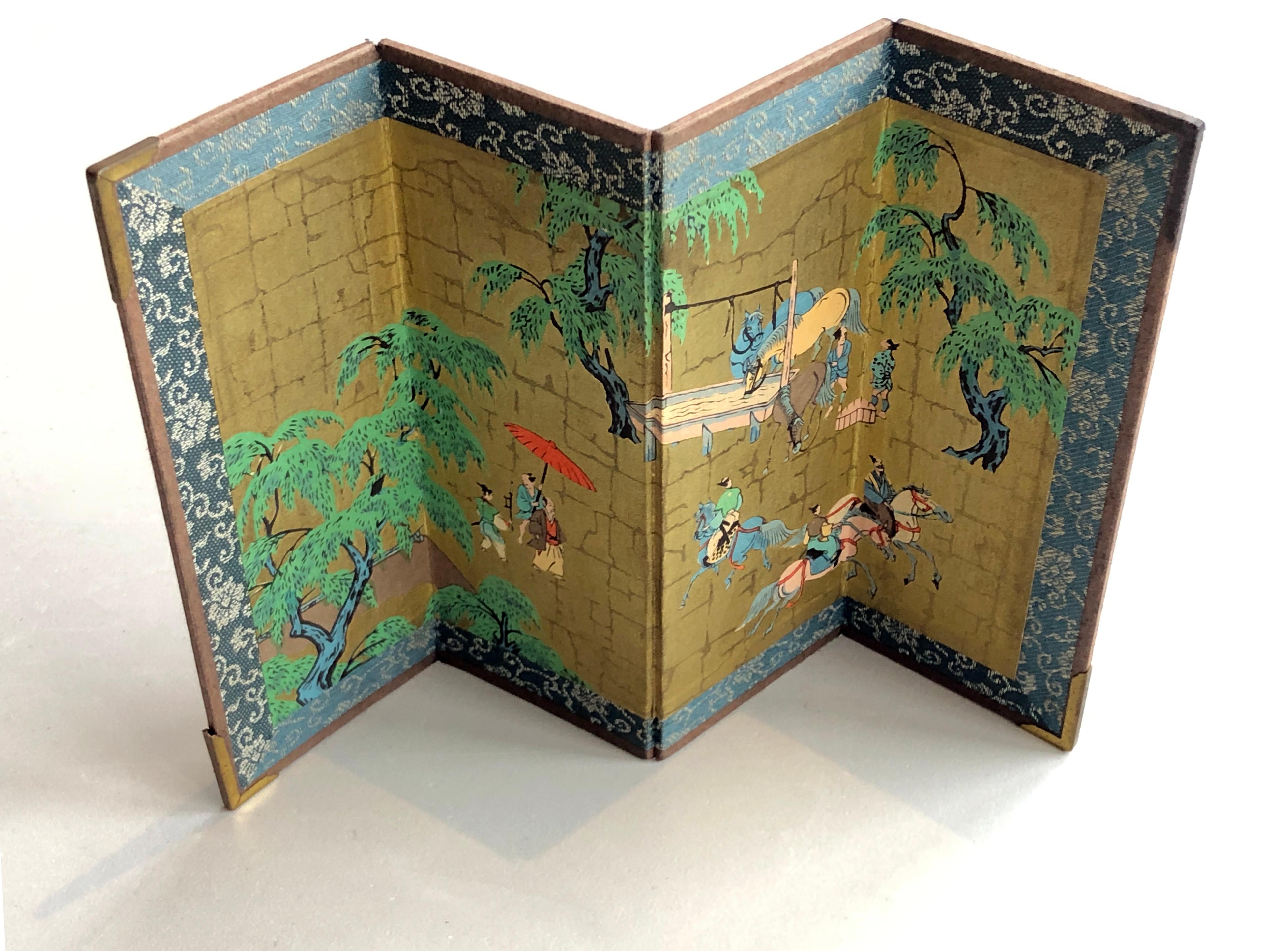 This is a Miniature Oriental Folding Screen featuring Asian painting artwork. The screen is adorned with gold tone corner protectors, although the top right corner one is missing. The background is patterned in gold with depictions of trees and