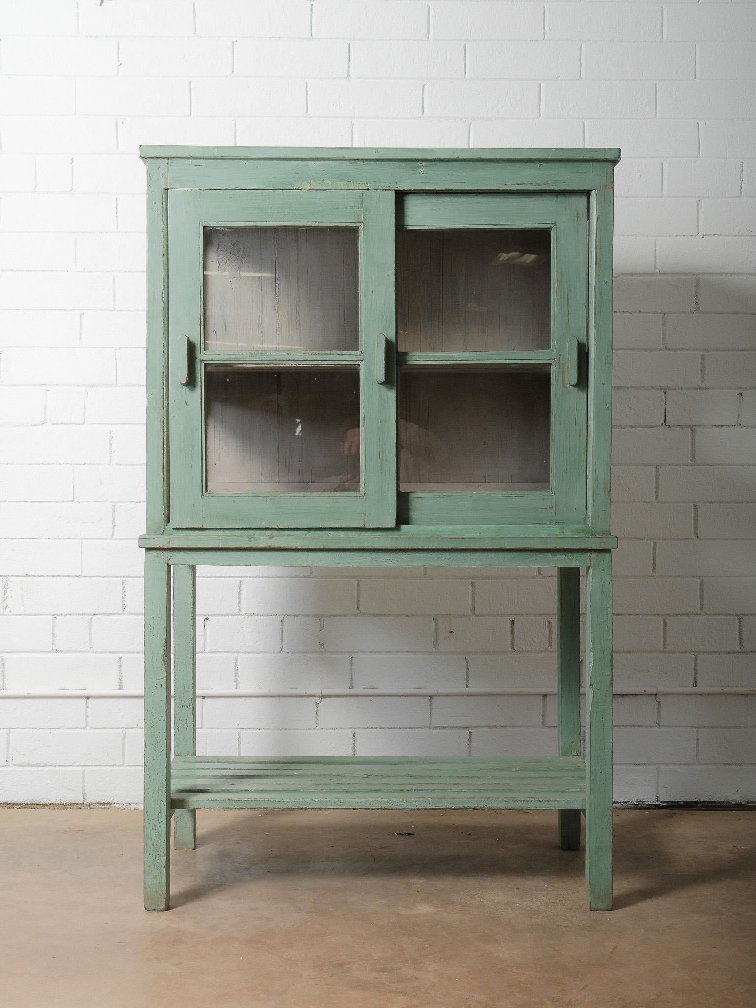 Une charmante armoire vintage en bois peint en vert menthe, avec une peinture joliment vieillie et des portes vitrées classiques. Reposant sur de hauts pieds avec une étagère inférieure ouverte, cette pièce allie des proportions légères et aérées à