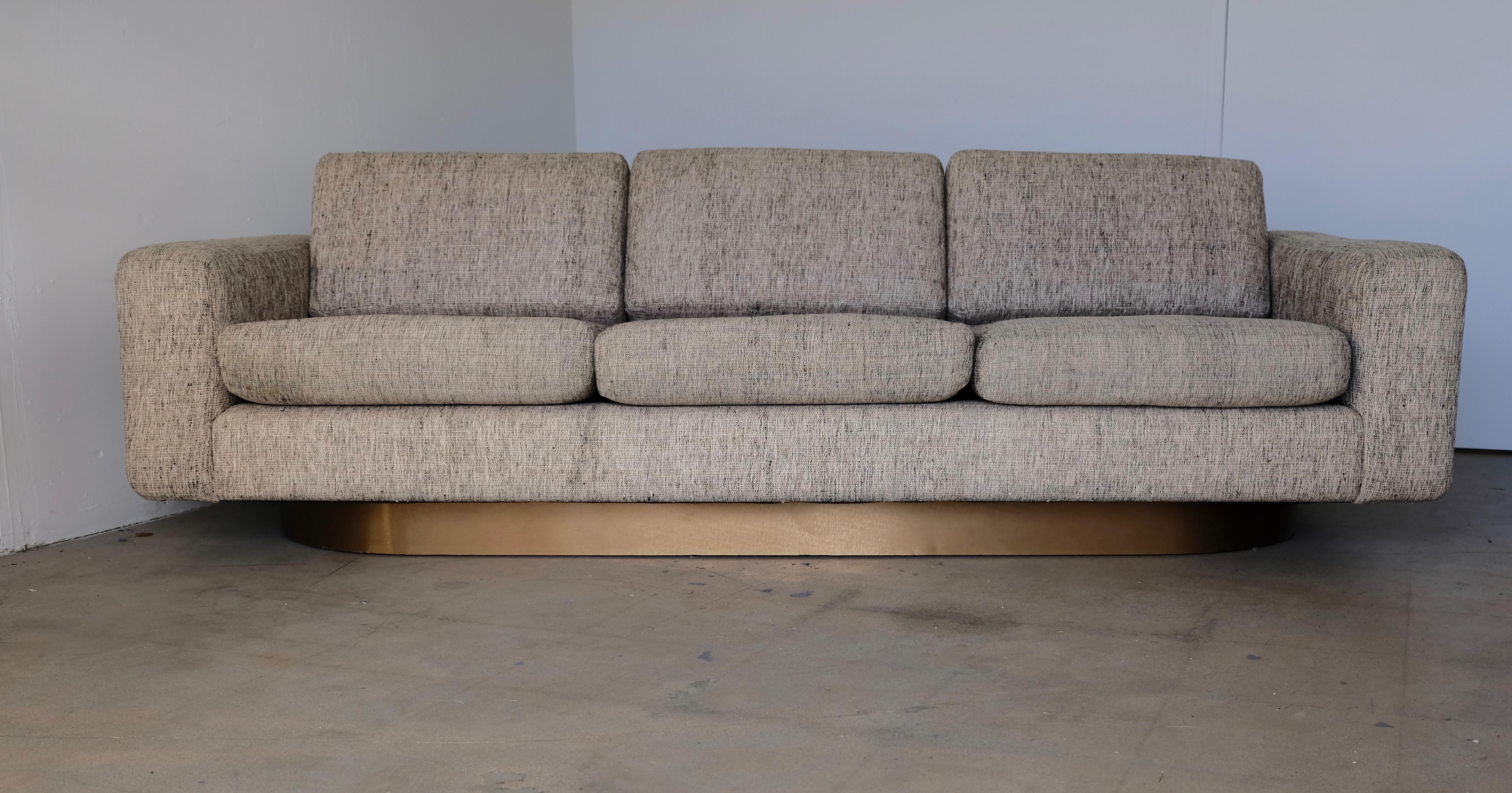 This custom modern three-seat couch has Classic midcentury lines. It floats on an oval shaped brushed brass covered base. The sofa retains its original tweed fabric which appears to be a silk blend. Measures: The arm height is 23