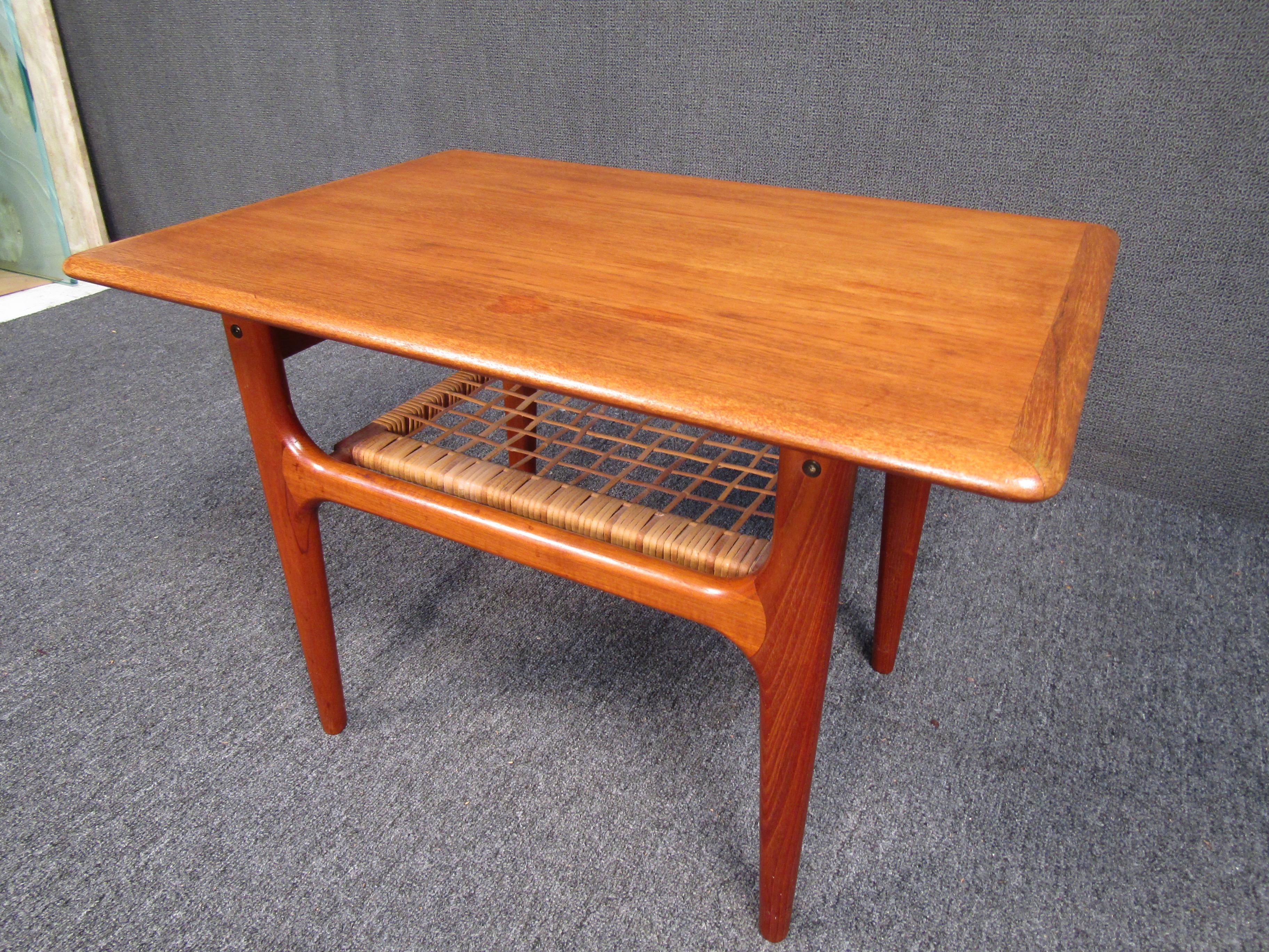 This fabulous Mid-Century Modern end table features a lower cane tier for added storage. The sleek design was made in Denmark and has elegant teak wood grain throughout. This lovely end table have smooth rounded edges and a rectangular top. A