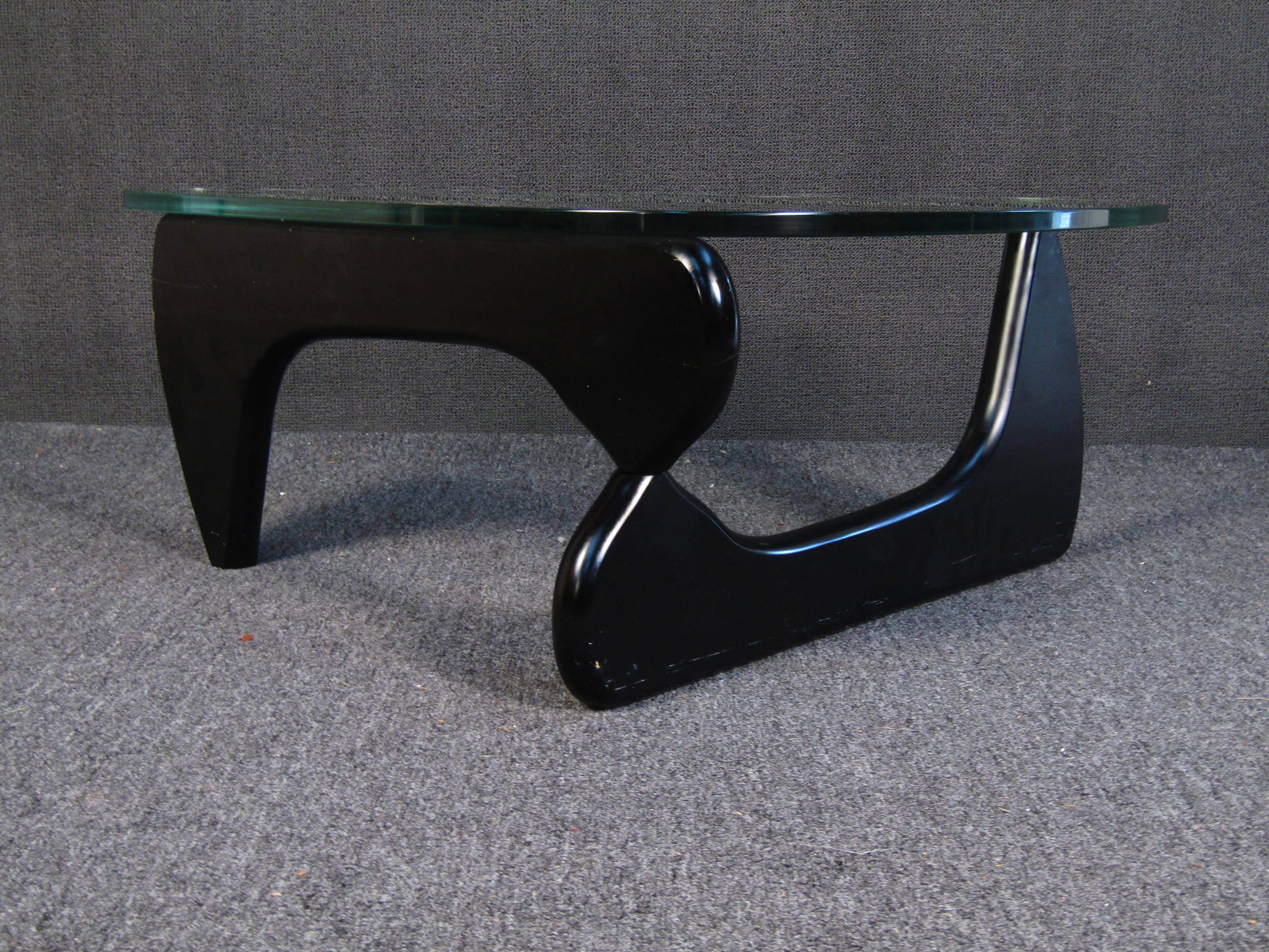 This Mid-Century Modern coffee table boasts two smoothly shaped pieces of solid wood. The pieces interlock in a tripod to support a thick glass tabletop. Please confirm item location (NY/NJ).