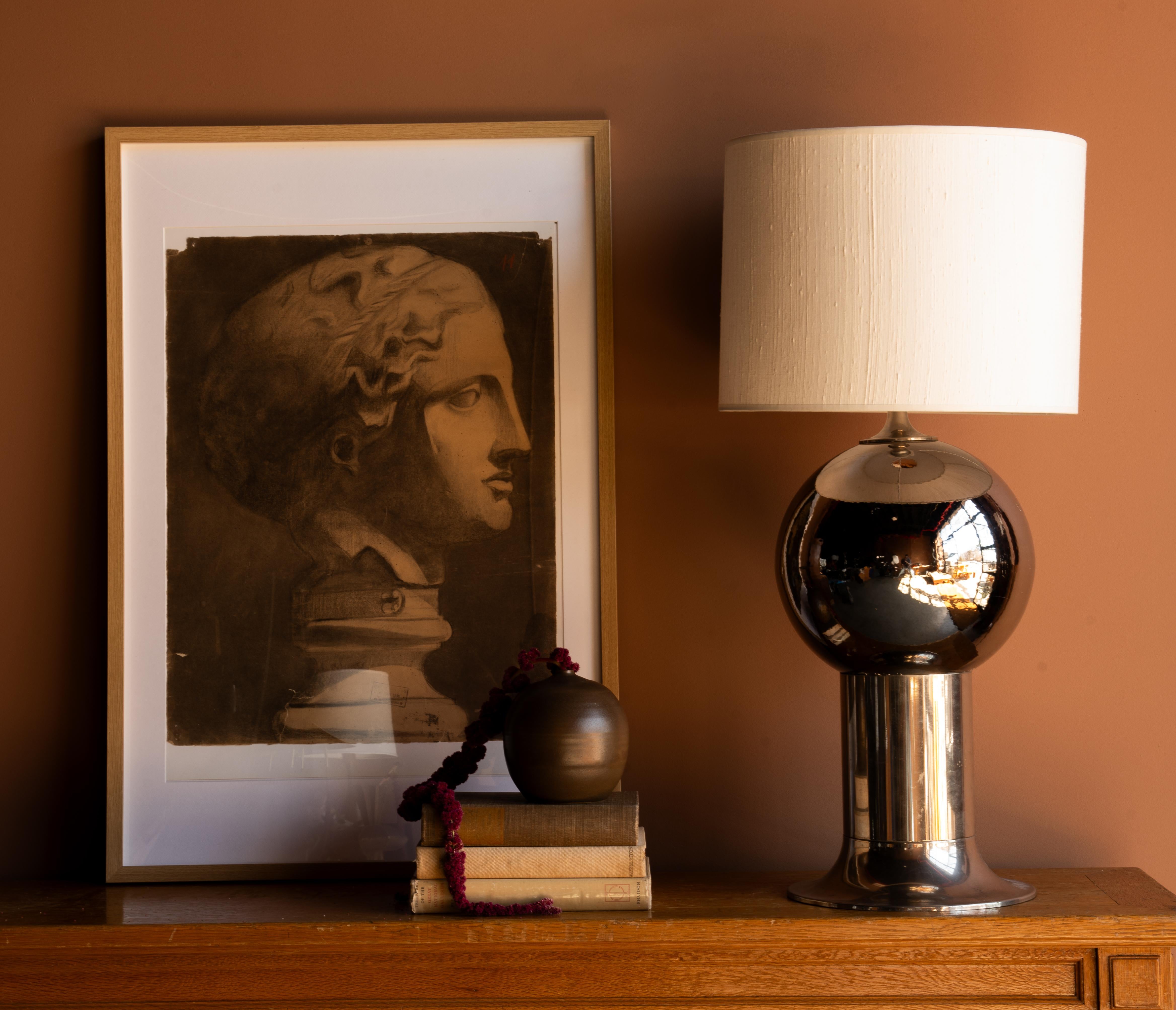 This lamp features a bold, sculptural design with a reflective chrome finish. The base consists of a cylindrical pedestal supporting a large, polished metallic sphere, creating a dynamic balance of forms. The included shade is textured off-white