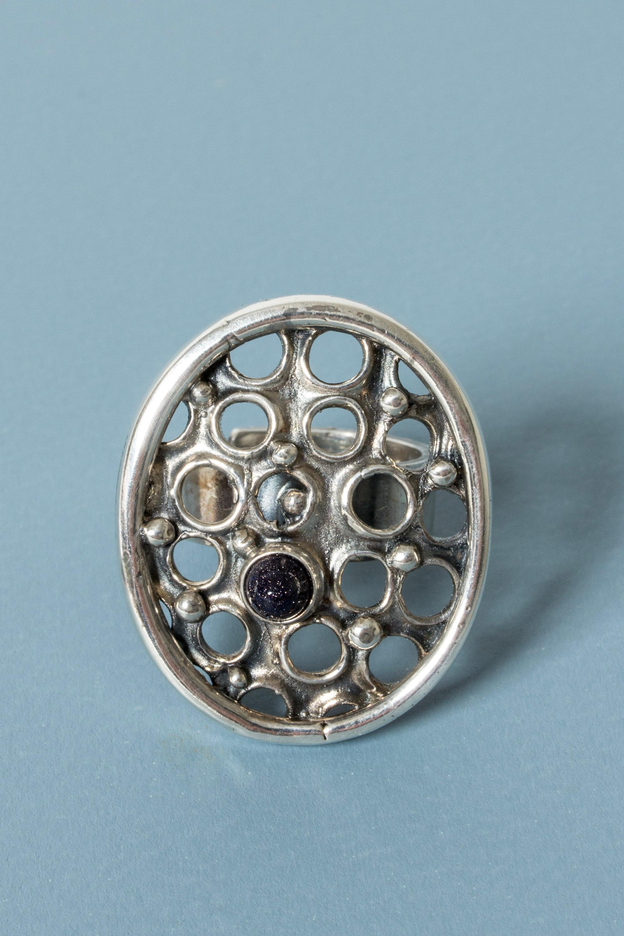 Expressive silver ring by Jorma Laine, with a large oval perforated with holes. A small bullet cut blue goldstone adds extra sparkle.

Jorma Laine was one of the most unique voices in the history of Scandinavian jewelry, and is considered one of the