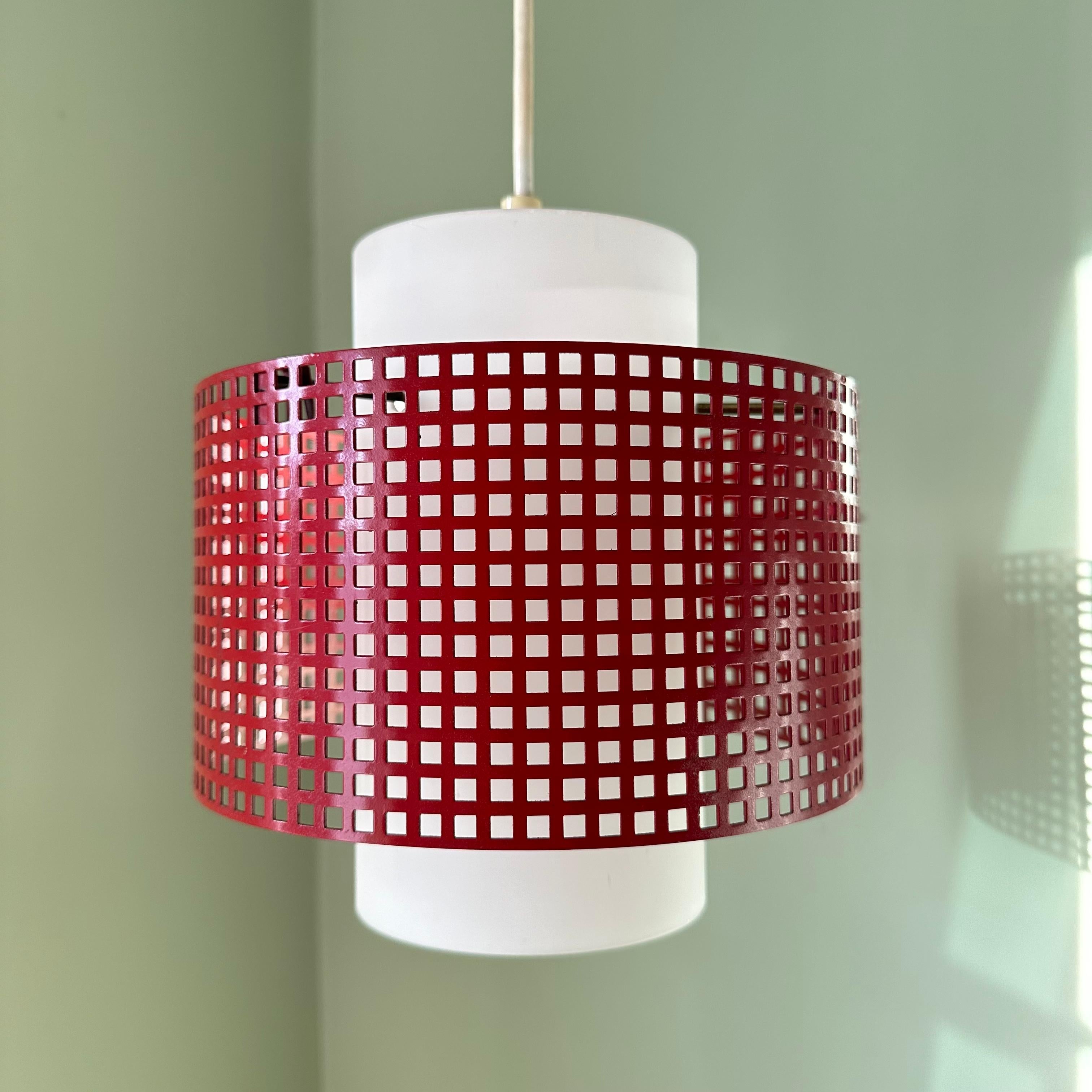 A vintage modernist or post-modern pendant ceiling light. A white glass cylinder diffuser shade floats inside a red painted metal ring, perforated into a grid or windowpane pattern. Suspended by brass rods, the red colored metal grid orbits around