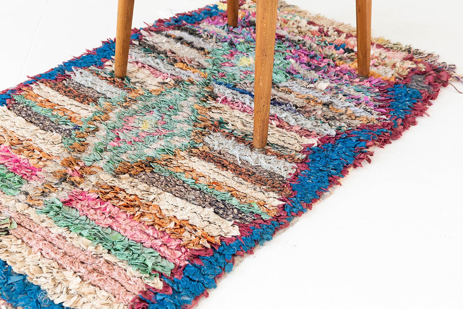 Streifen, Zickzacklinien und zentrale Rautenmotive in einer lebhaften Farbpalette aus leuchtendem Blau, Burgunderrot, Apricot, hellem Goldgelb und Weiß. Der Flor besteht aus bandartigen, recycelten Textilien. Ein fröhlicher Vintage-Bandteppich aus