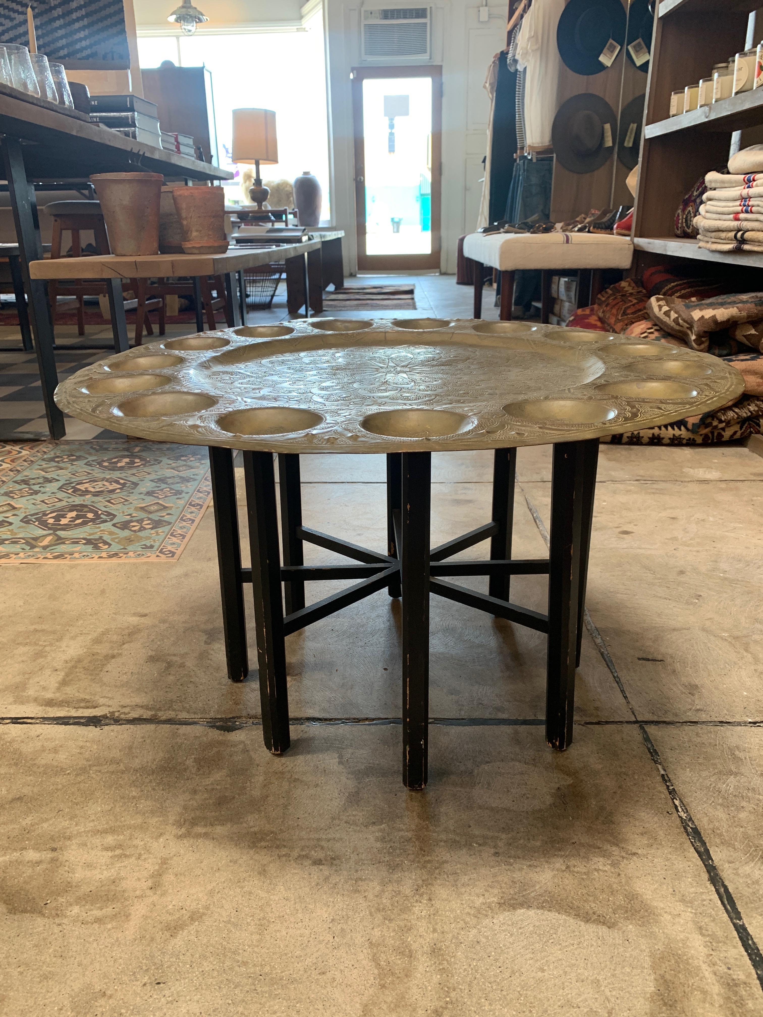 Vintage brass tray table from Morocco, features indents around the circumference of the tray for cups or glasses, and intricate designs carved into the metal. Accompanied by a wooden Stand painted black. This table is great as a side or end table,
