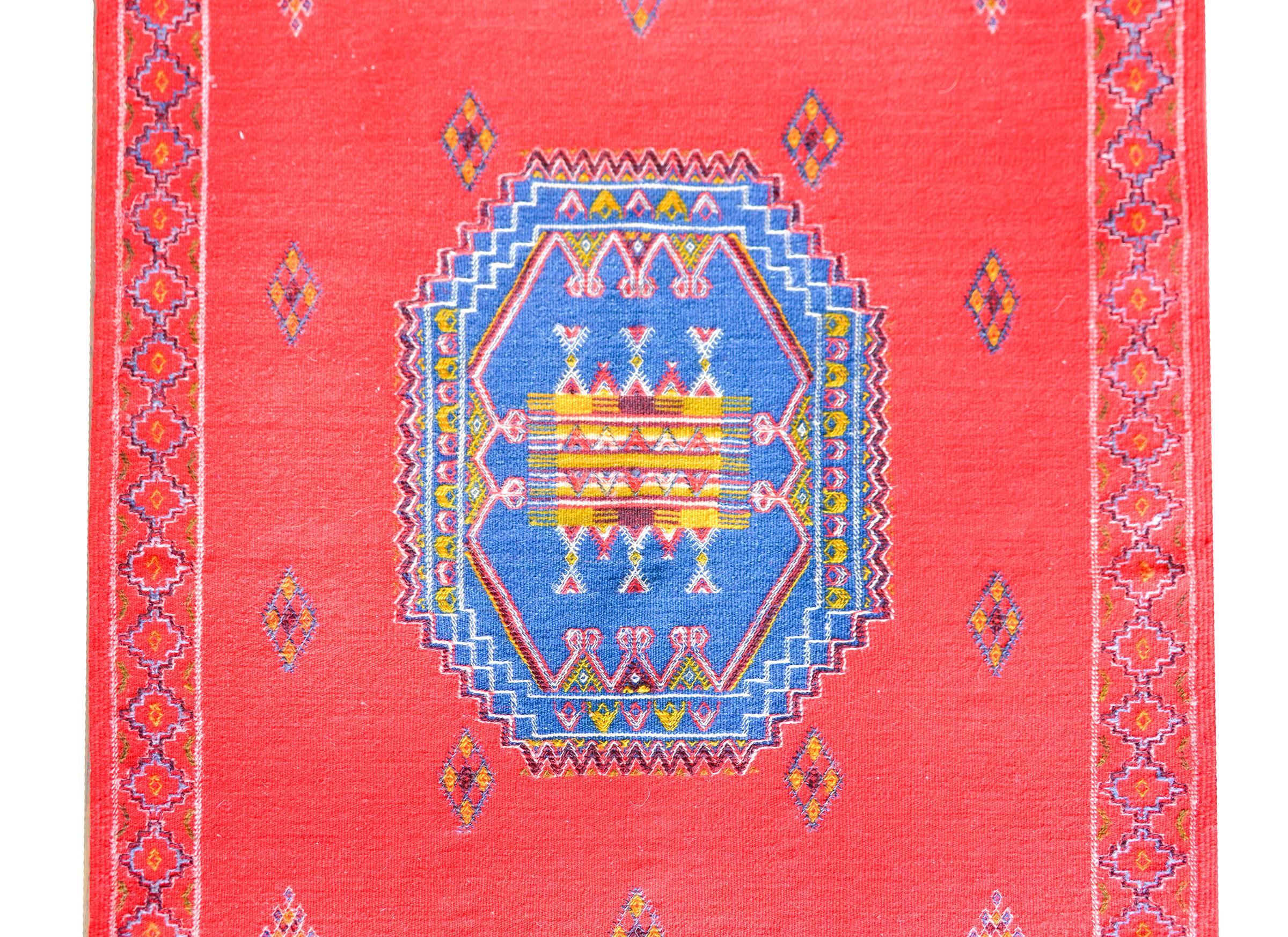 A bold Moroccan flatware rug with three large medallions with stylized floral patterns against a crimson field with more stylized flowers. The border is simple with an embroidered geometric pattern.