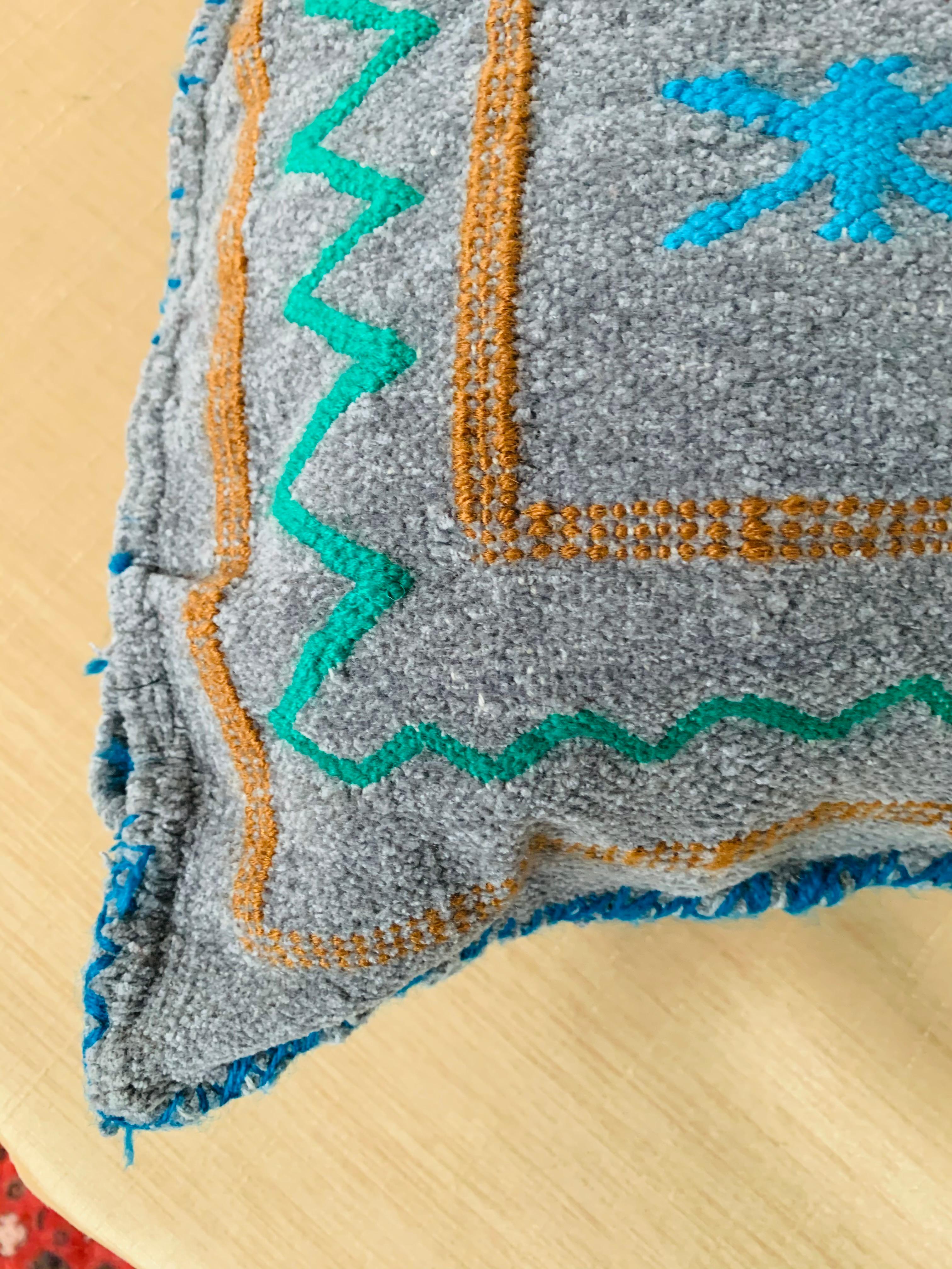 Ein einzigartiges, handgewebtes marokkanisches Kelim-Kissen aus weicher Wolle mit mehrfarbigem Berberstamm-Muster auf grauem/blauem Hintergrund. Es besteht aus einer Mischung aus weicher Schafwolle und natürlichen organischen Farbstoffen.

