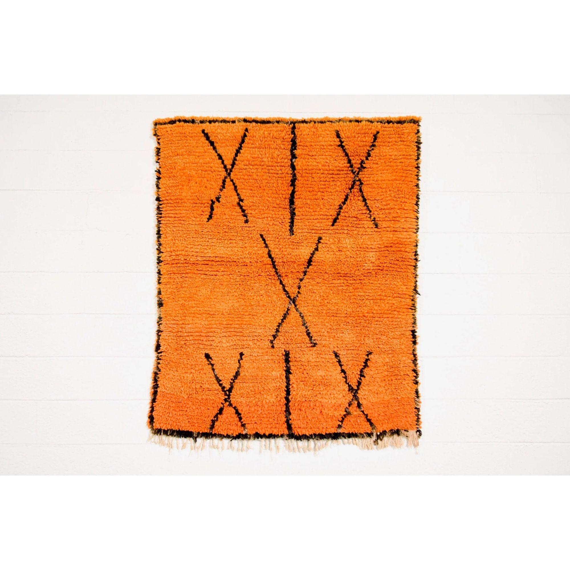 This vintage handwoven Moroccan Azilal tribal floor rug features a design of asymmetrical black crisscross shapes and border edge set against a stunning tangerine orange color field. The thick wool pile is shaggy and full and the area rug is