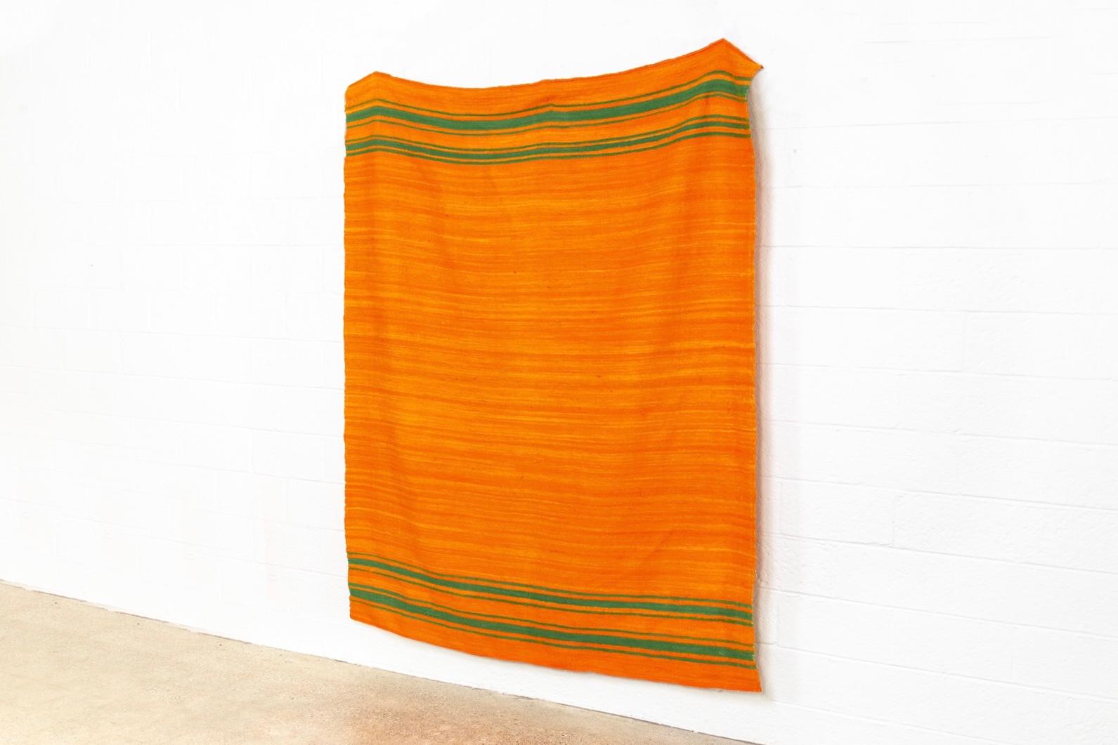 This vintage handwoven Moroccan Berber Kilim rug is handcrafted from hand-spun wool. It features a beautiful tangerine orange field with green stripes at both ends. The Minimalist design pairs well with a variety of decor styles including