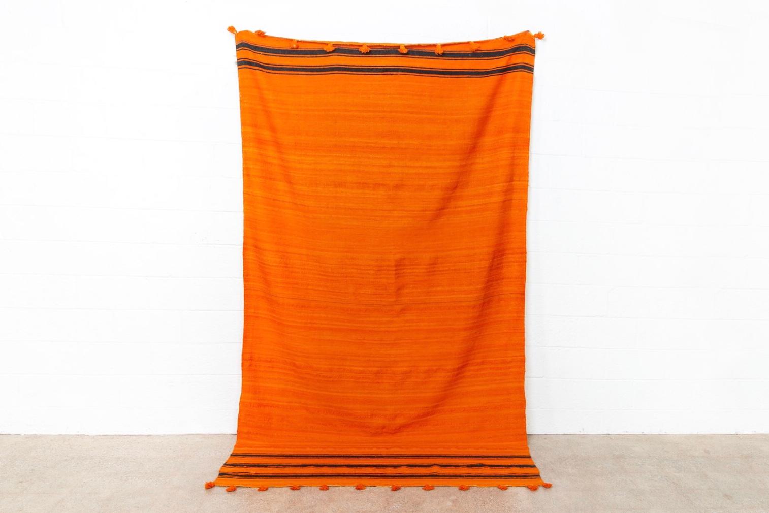 This large vintage handwoven Moroccan Berber Kilim rug is expertly handcrafted from hand-spun wool. The stunning tangerine orange field has subtle variation in shade and features double and triple black horizontal stripes at either end. The rug is