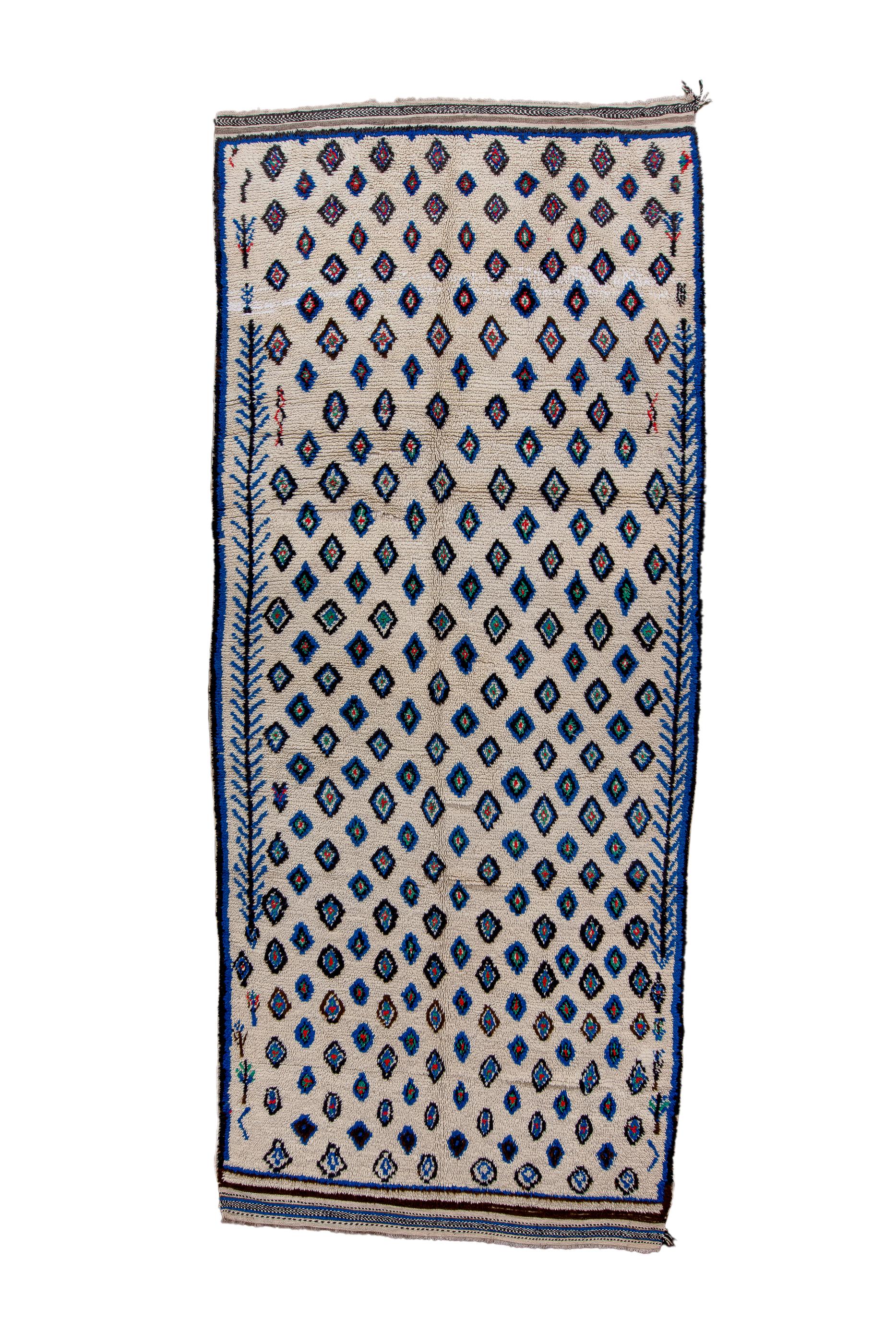 The ecru field shows a repeating, offset pattern of irregular, charming small lozenges, forming an offset lattice, with long herringbones up the sides. Striped flatwoven finishes at each end.  Blue and dark brown accents overall. Semi-vegetal shapes