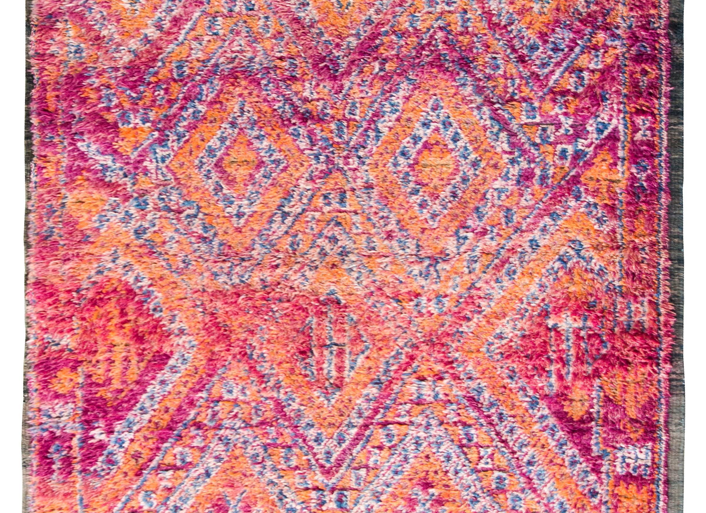 A bold and beatiful vintage Moroccan rug with a repeated diamond pattern surrounded by a repeated geometric patterned border, and all woven in bright oranges, fuchsias, and violets.