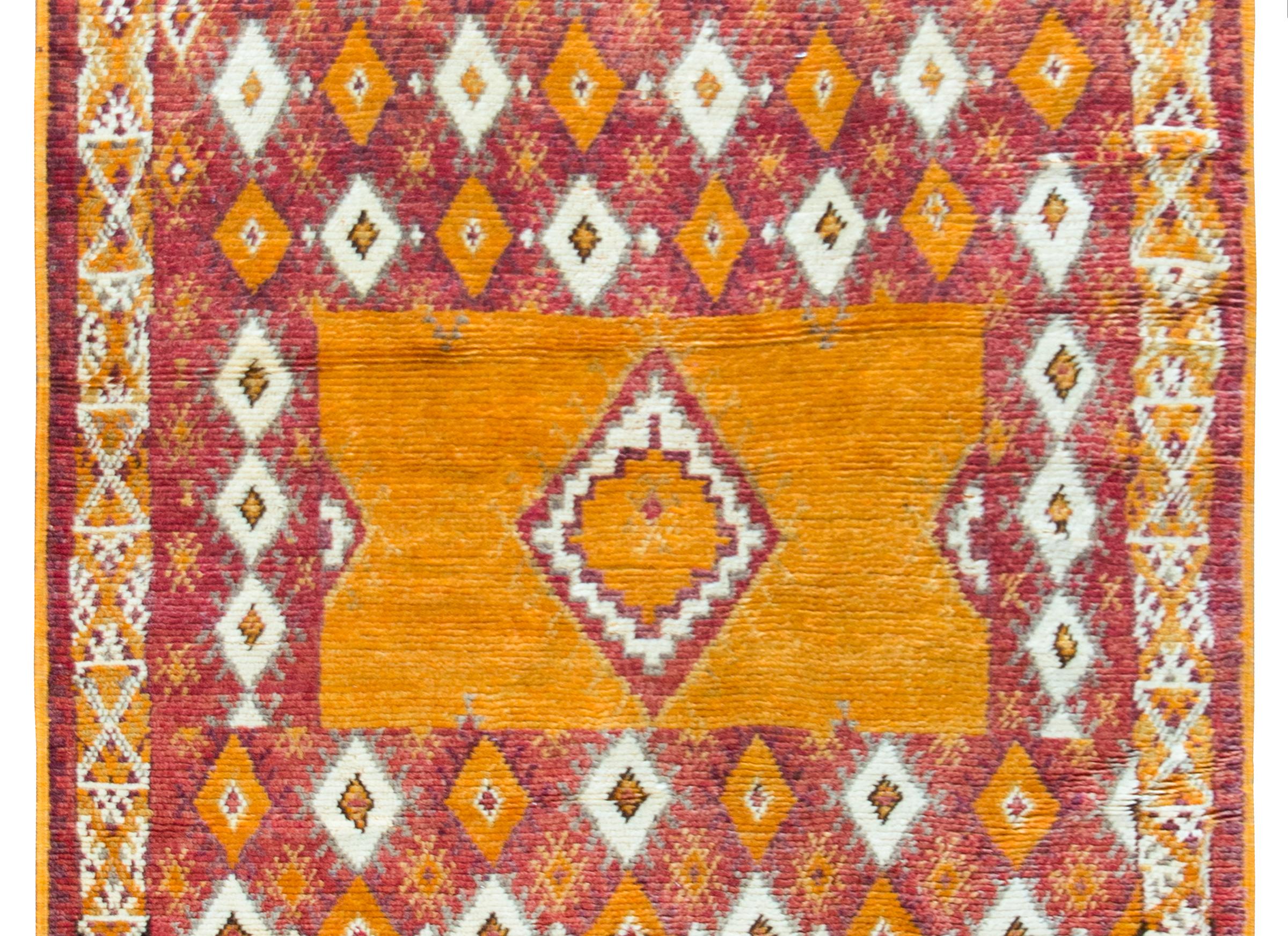 A bold vintage mid-20th century Moroccan rug with a large central diamond medallion set against an orange rectangular ground, and all set against a repeated cranberry, violet, orange, mustard and cream colored diamond patterned background, and all