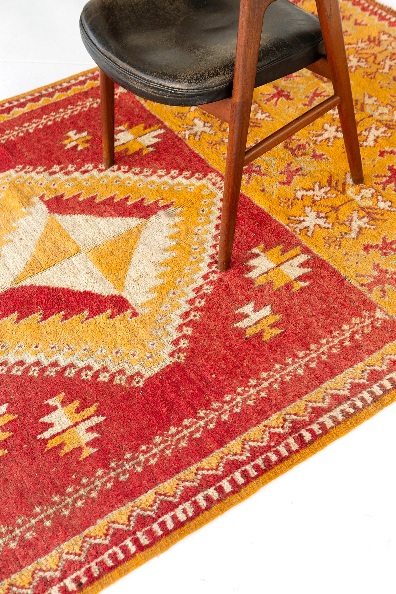 Au centre de la scène se trouve un médaillon losange vibrant qui s'étend sur les motifs floraux ornés de ce tapis marocain vintage inspiré de la tribu du Haut Atlas. Son ambiance vibrante, caractérisée par des nuances de jaune canari et d'orange,