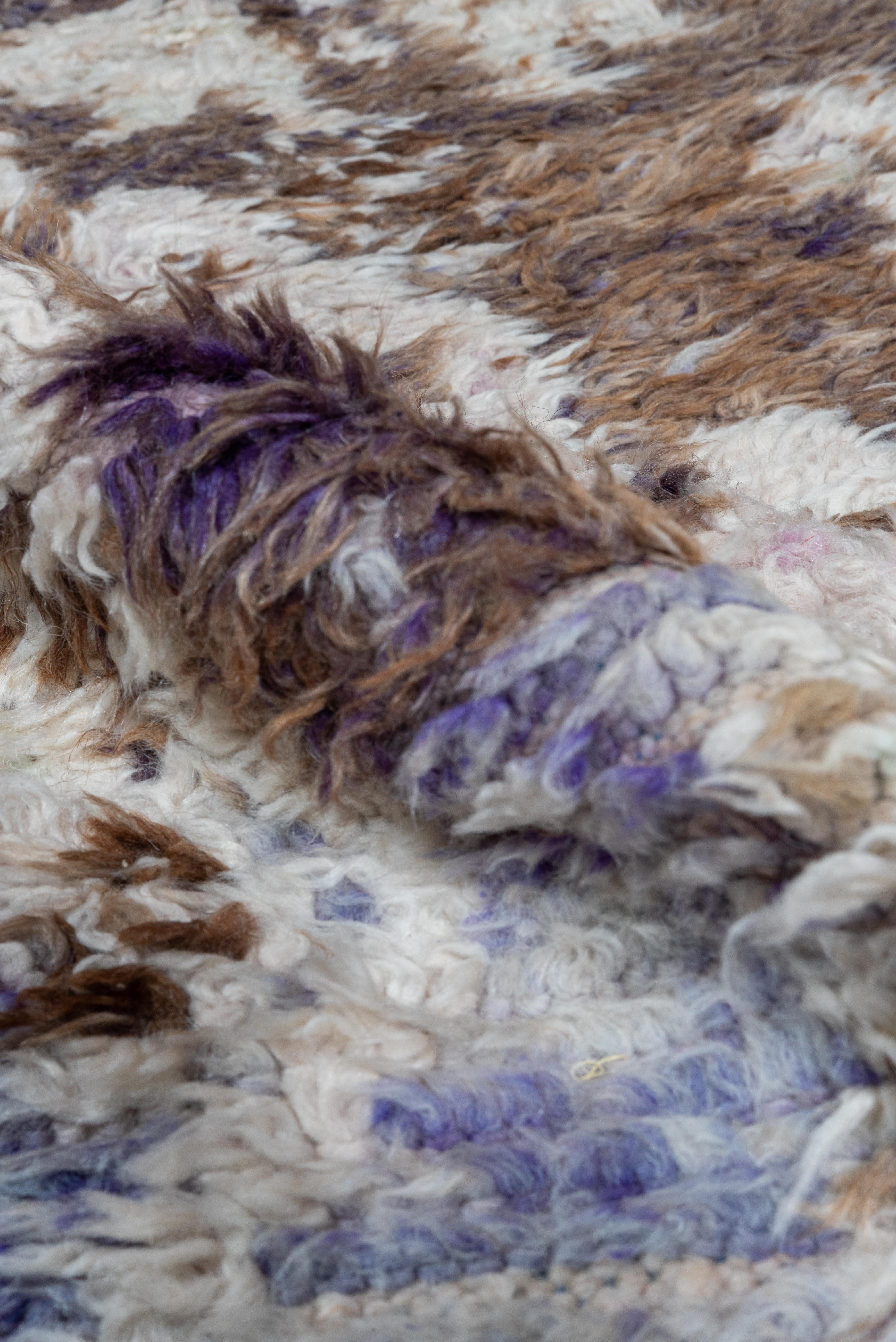 Una alfombra marroquí vintage ricamente estampada que se distingue por su suave paleta neutra de marrones terrosos, beige cremoso y tenues matices lavanda. De la superficie afelpada emergen intrincados motivos tribales y formas de medallones,