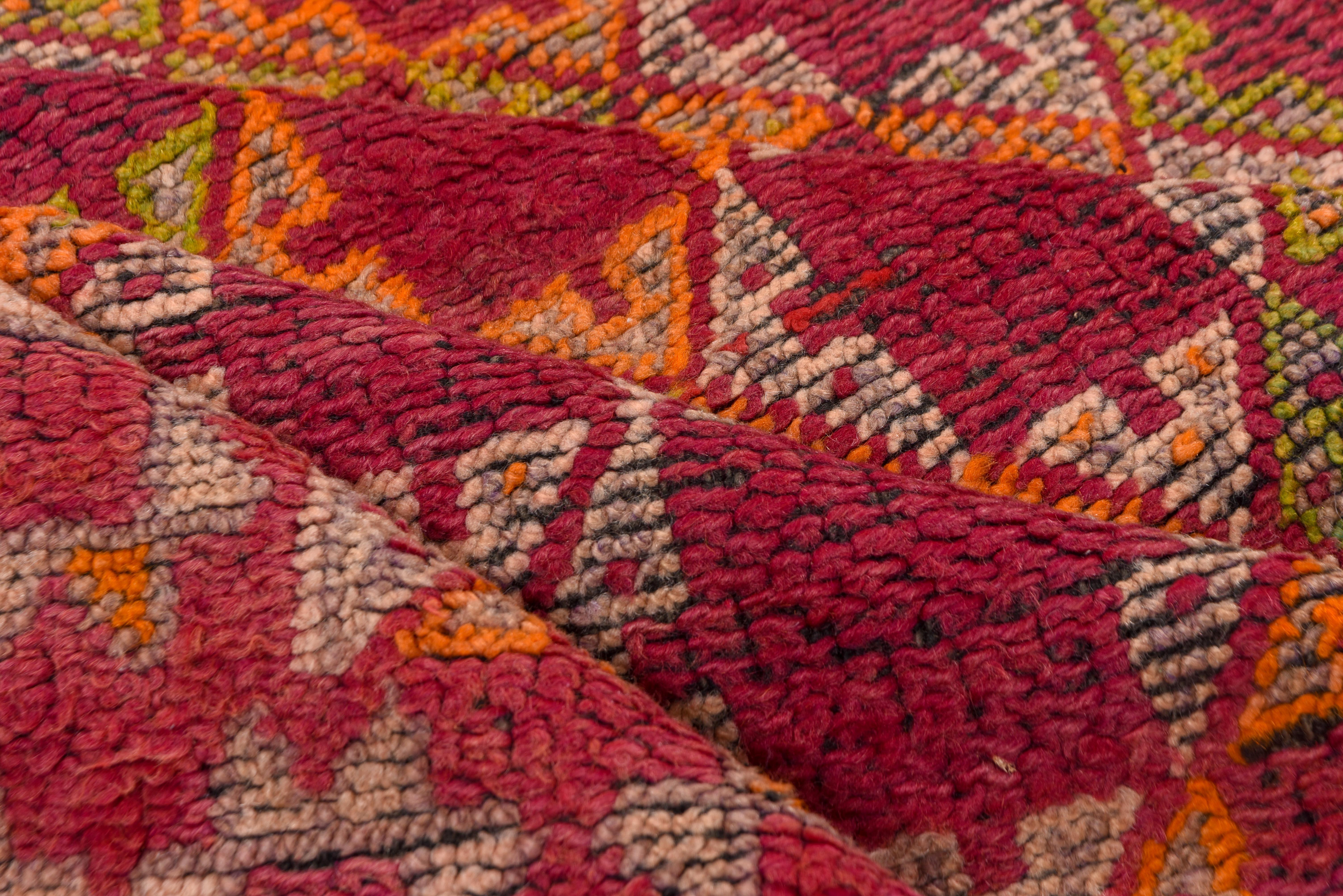 Questo tappeto marocchino vintage presenta un sorprendente reticolo di motivi a stella a forma di X nei colori rosso intenso, verde oliva, zafferano dorato e avorio, incorniciato da un bordo geometrico di rosette stilizzate. Il fondo cremisi