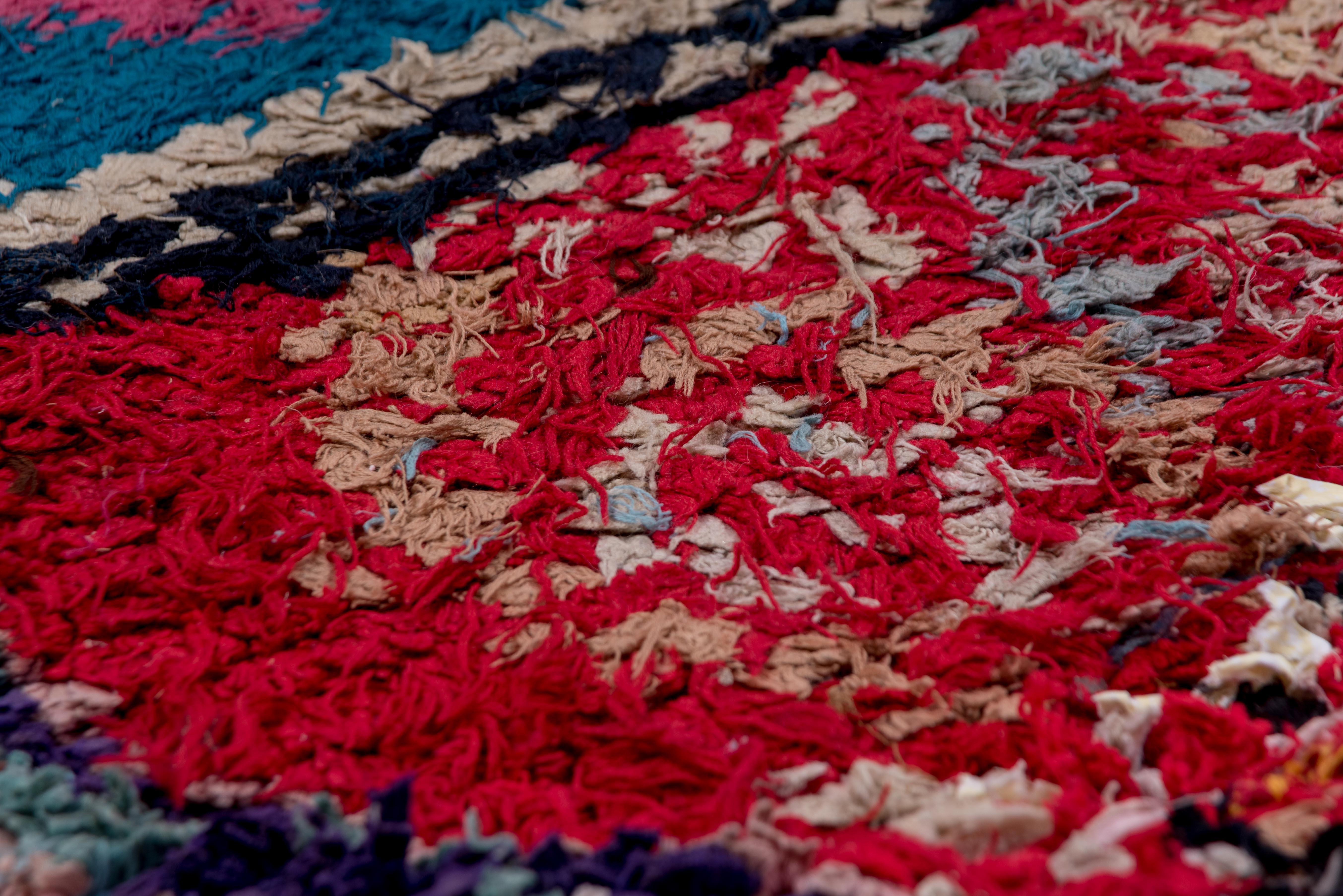 Una alfombra marroquí audaz y cautivadora que se distingue por su campo carmesí saturado y un animado conjunto de motivos tribales en forma de rombo. La pieza rebosa vitalidad cromática, con tonos azul cobalto, fucsia, azafrán y crema que se