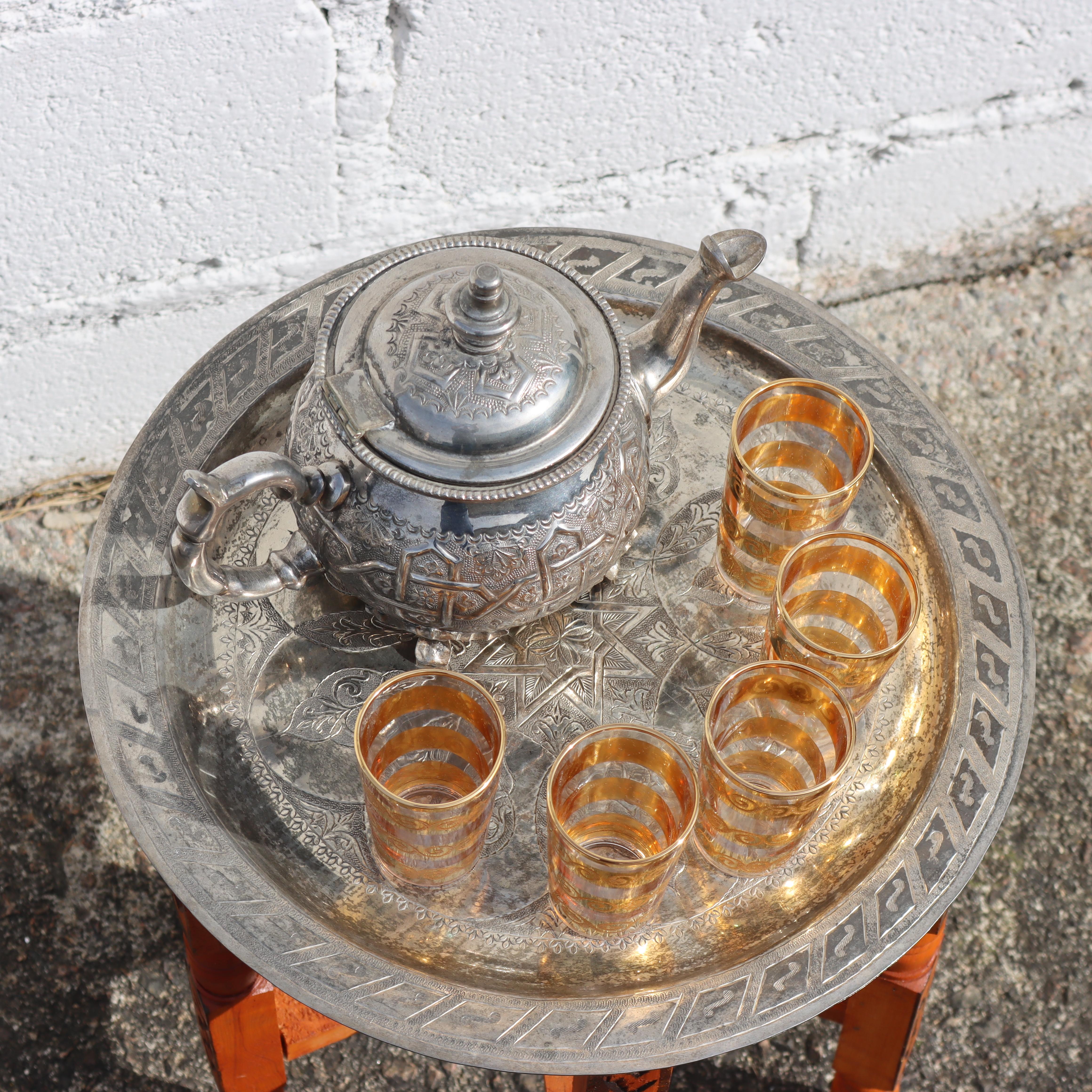 This exquisite Moroccan tea ceremony table from the 1980s features an ornate, solid tabletop crafted from silver-plated brass, adorned with intricately etched motifs. 
The plate rests upon a beautifully carved folding base with six legs, each