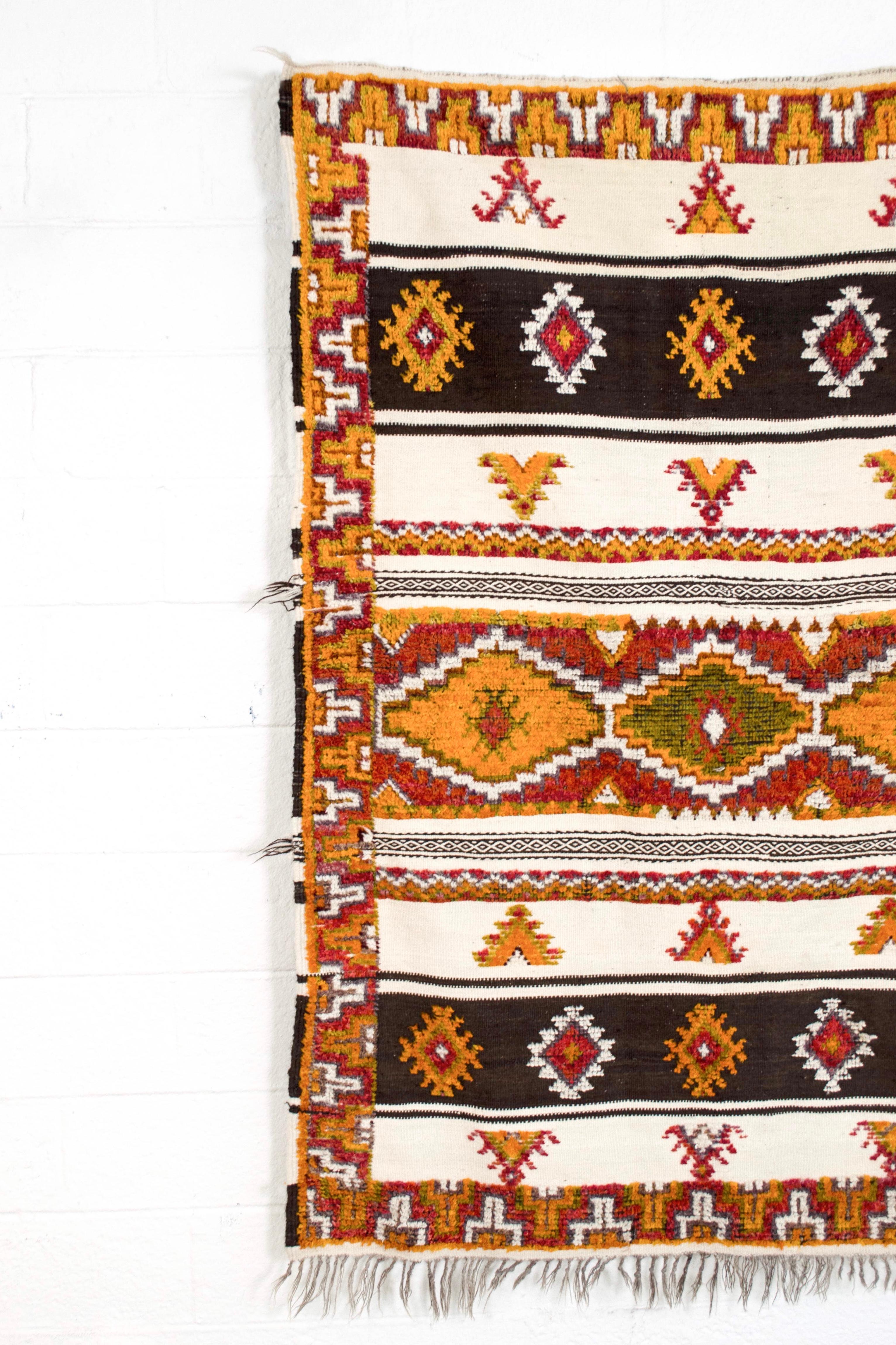 This beautiful vintage handwoven Moroccan Berber rug is circa mid-20th century. It features a symmetrical banded geometric design infused with tribal symbolism in gorgeous shades of orange, red, pink, green, gray, brown and cream with gray fringe to