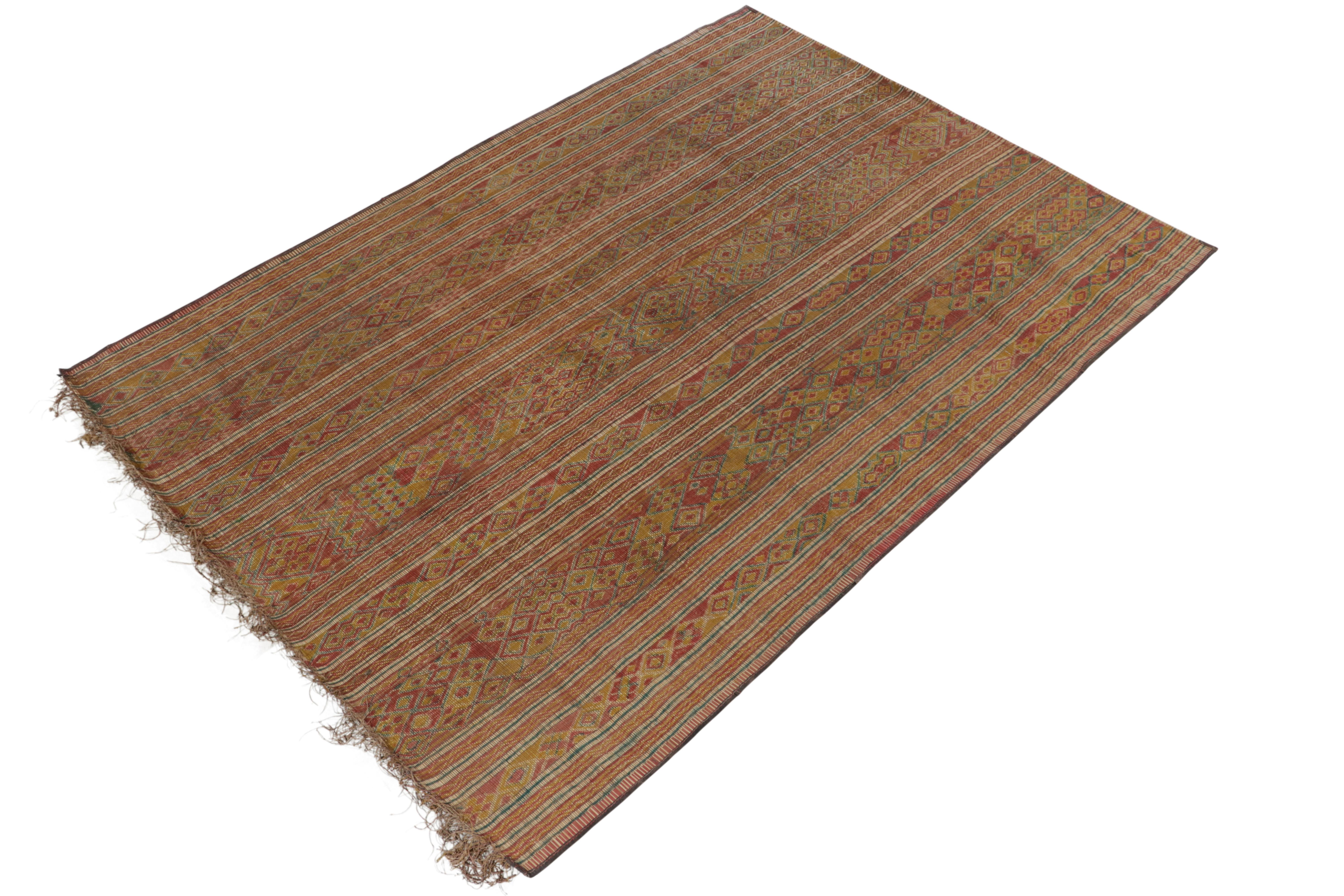 This vintage Moroccan rug is an 8×10 Tuareg mat from the nomadic tribe of the same name. Handwoven with reeds and leather circa 1950-1960, its natural materials enjoy warm beige-brown undertones to gold, green and red stripes and geometric