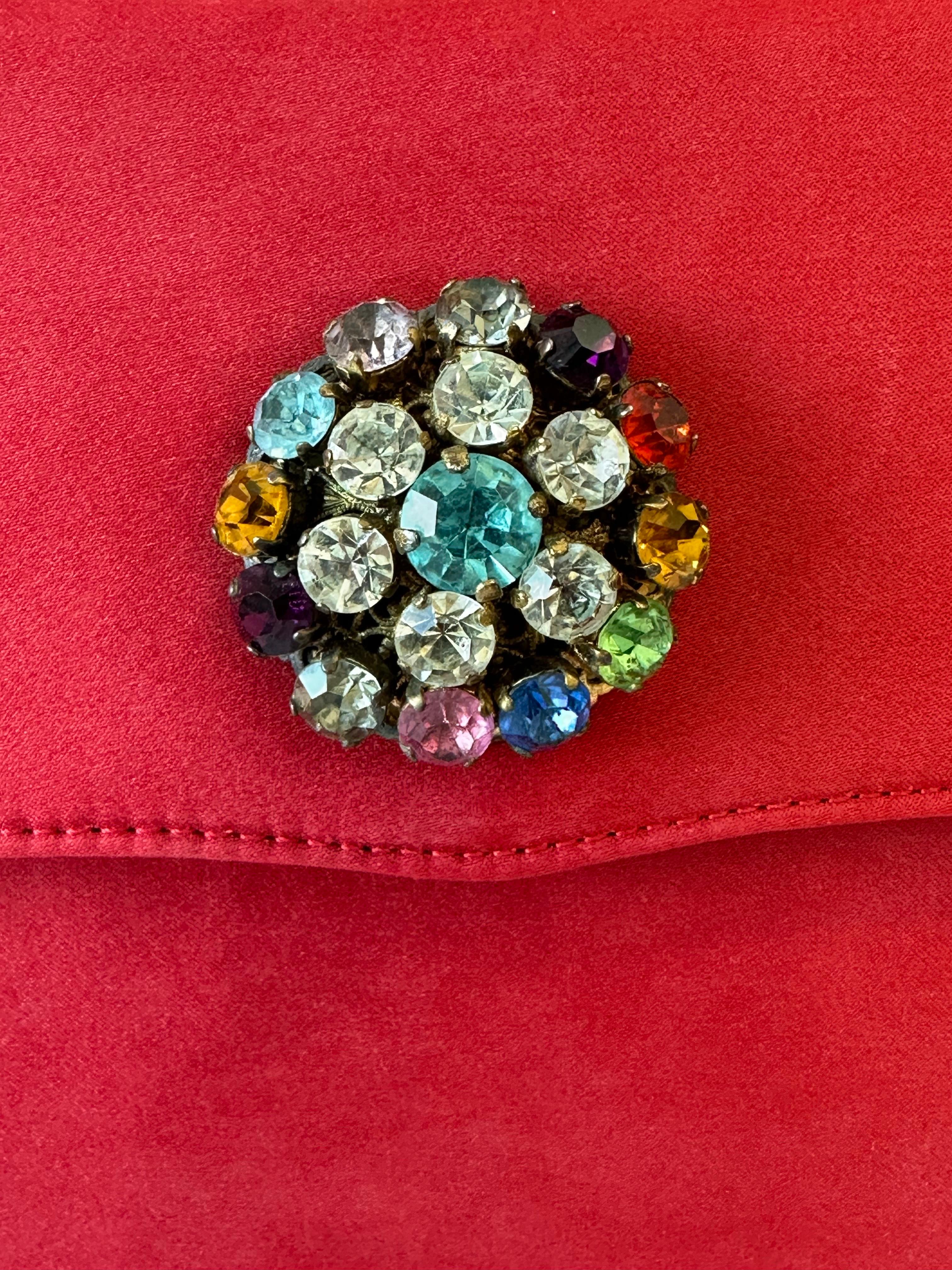 Vintage Moschino
Redwall satin flap handbag.
Secure magnetic closure adorned with a striking multicolored stone encrusted round jewel. Black canvas interior with a spacious main compartment and a zippered pocket featuring the iconic Moschino