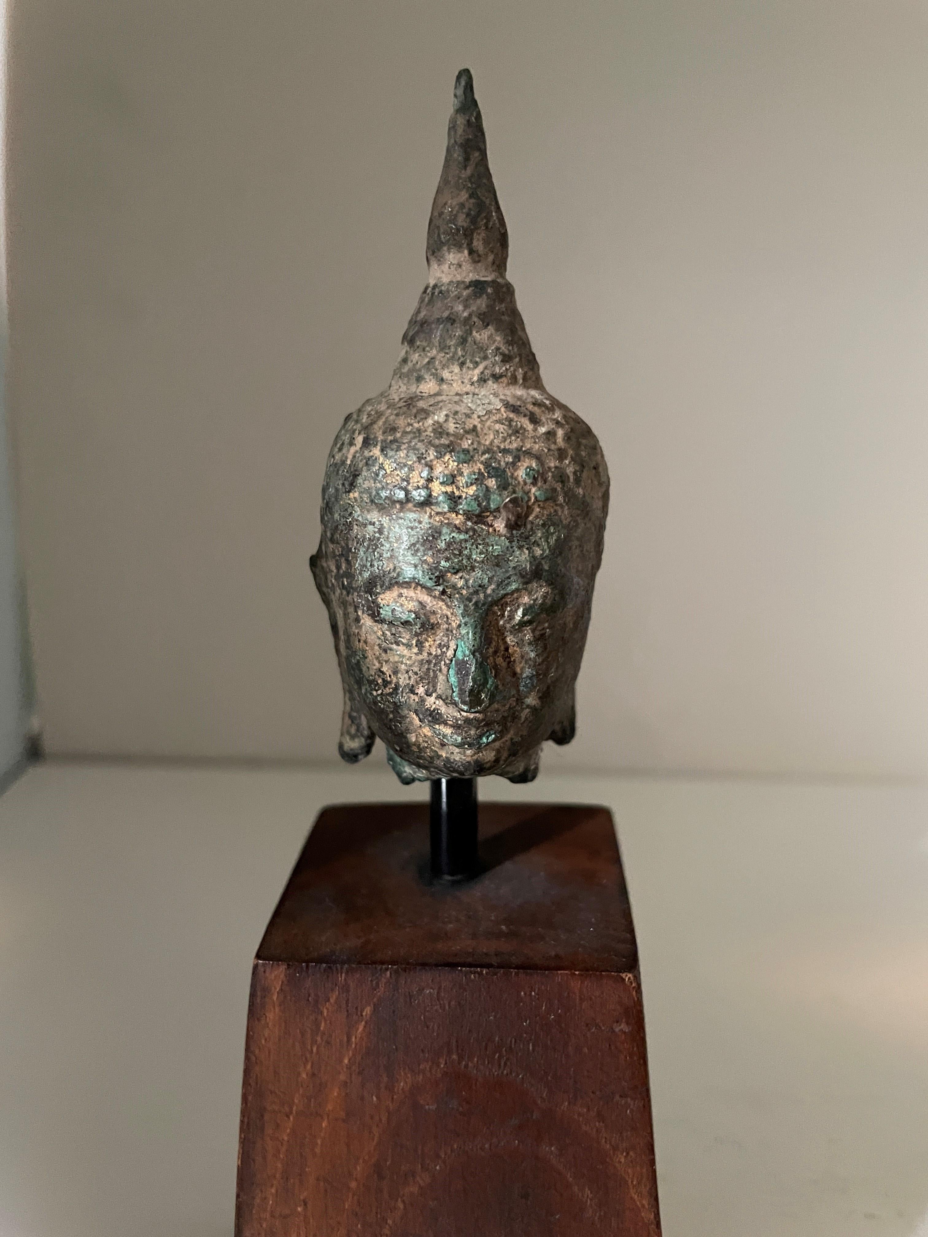Kleine antike Bronzebüste des Buddha-Kopfes auf massivem Holzsockel. Der Kopf ist stark in Mitleidenschaft gezogen und patiniert, was auf ein hohes Alter schließen lässt; die Rückseite des Kopfes weist einige kleinere Schäden auf, die aber nichts
