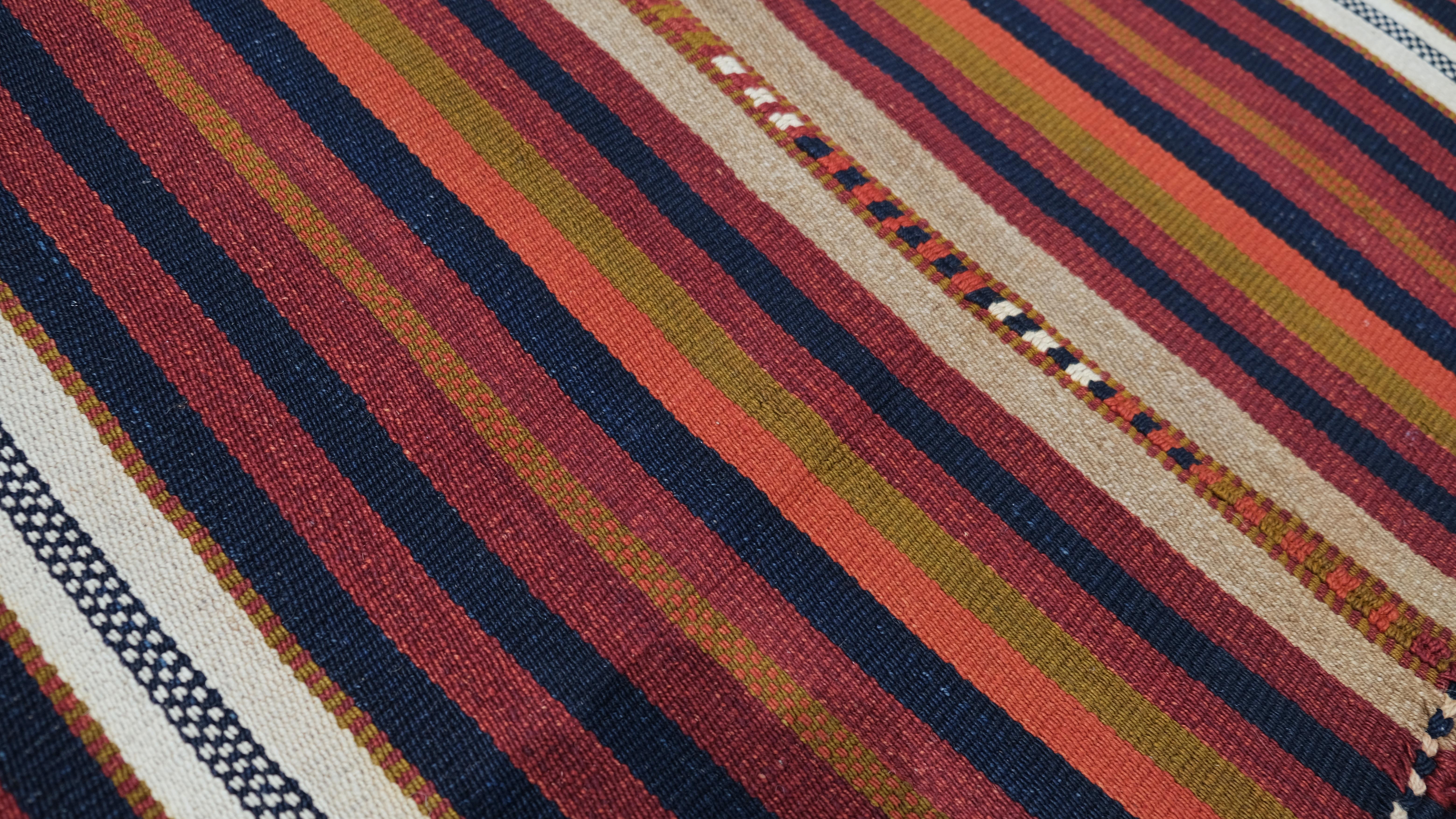 Dieser handgewebte Kelimläufer aus Wolle ist ein atemberaubendes Beispiel traditioneller Handwerkskunst mit leuchtenden, mehrfarbigen Streifen in satten Erdtönen. Die abwechselnden Farbtöne von tiefem Rot, Marineblau, Orange und Vory bilden eine