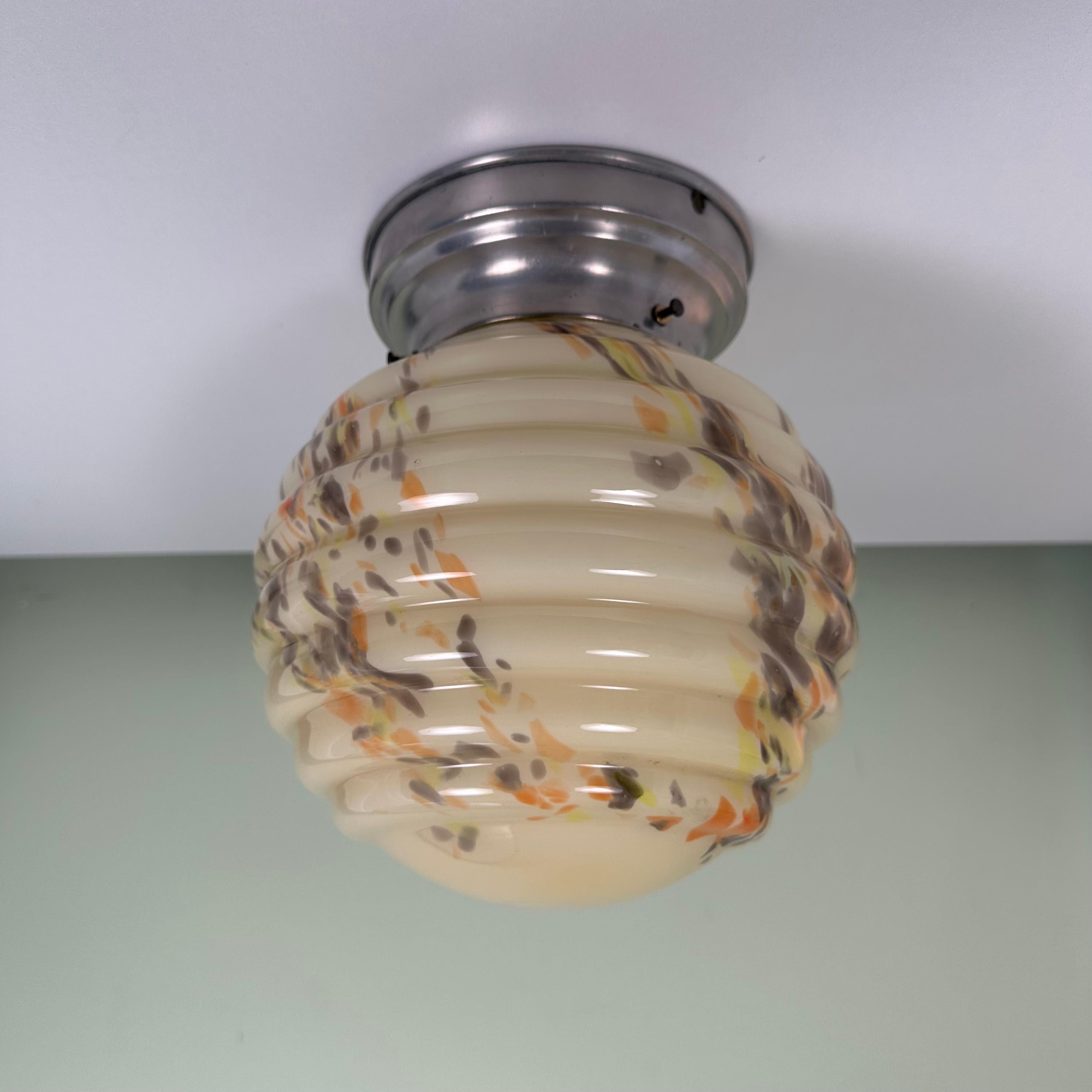 Vintage glass globe flush mount light. The perfect midcentury color scheme meets an Art Deco Shape. A cream colored base with tie-dye-esque colorful inclusions. The colors are orange, neon yellow, and smoke gray. As these colors overlap, slight