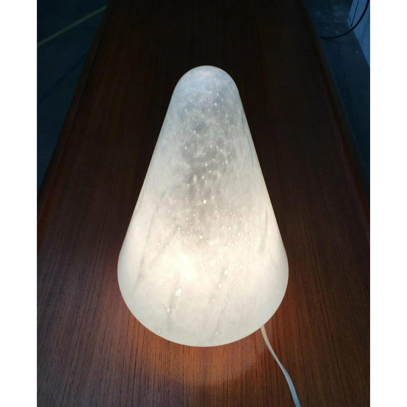Cone table lamp made of Murano glass.

The cone-shaped diffuser is made entirely of glass. The glass shows a wild game of inclusions, bubbles and gradients. The shape of the cone tapers at the bottom and appears to be above the ground. The frame