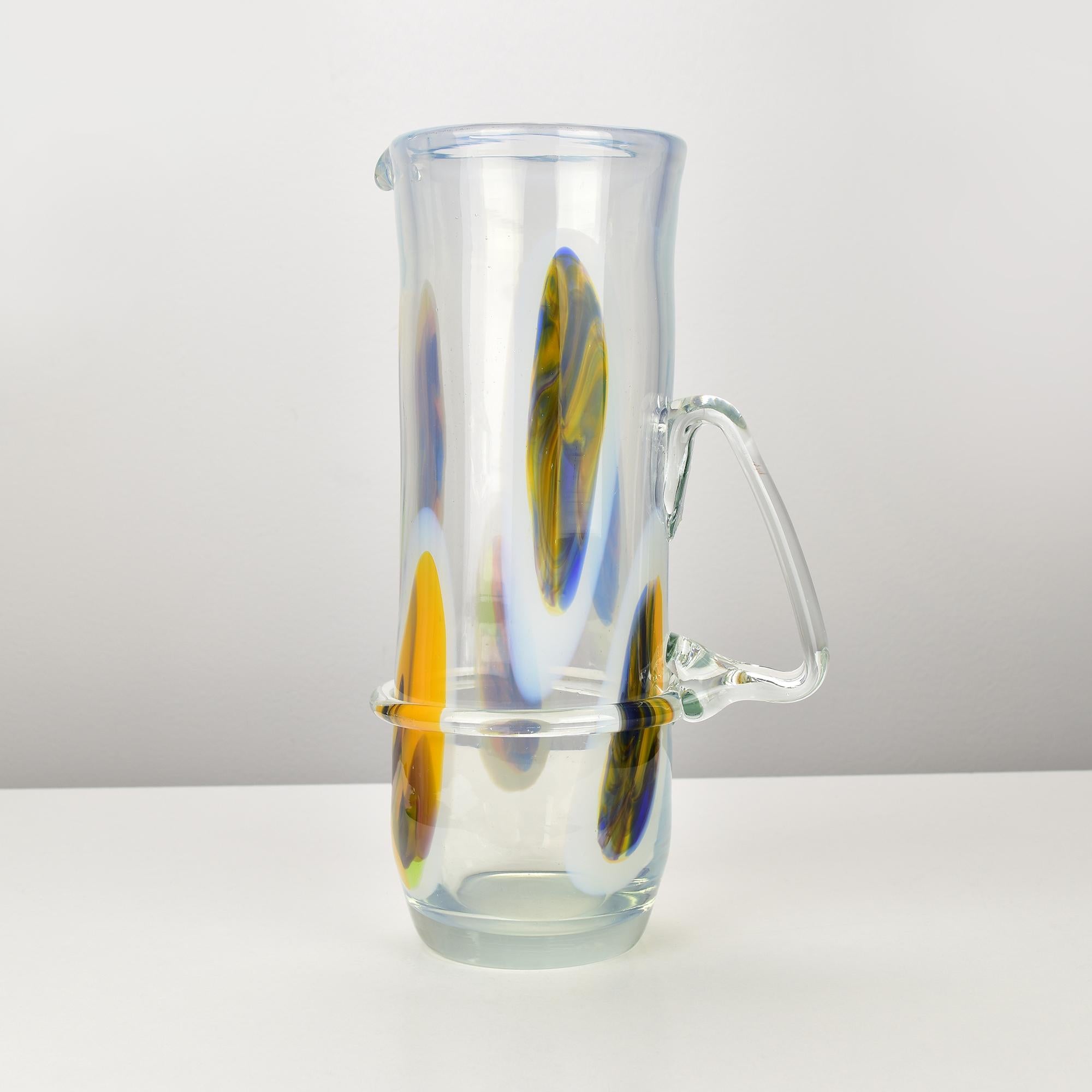 Questa brocca vintage in vetro di Murano esemplifica l'arte italiana con i suoi caratteristici accenti colorati in giallo e blu, racchiusi in un vetro trasparente. Il pezzo è caratterizzato da disegni ovali allungati che danno un tocco vivace e