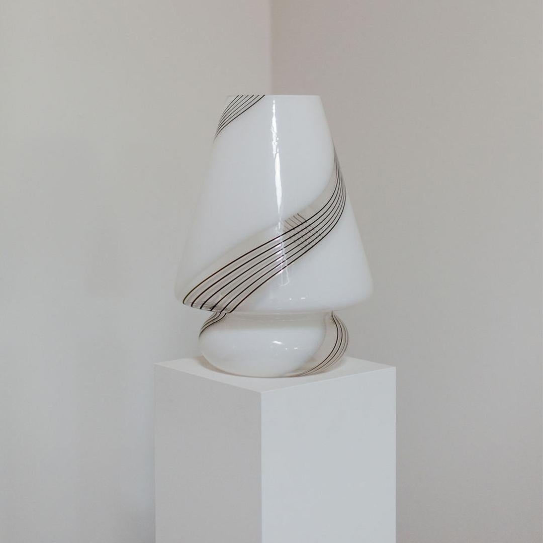 Lampe de table à champignon en verre de Murano laiteux soufflé à la main, datant des années 1960. Fabriquée avec le plus grand art, cette lampe incarne l'essence du rétro chic avec sa silhouette gracieuse et ses rayures grises et noires. 

Câblée