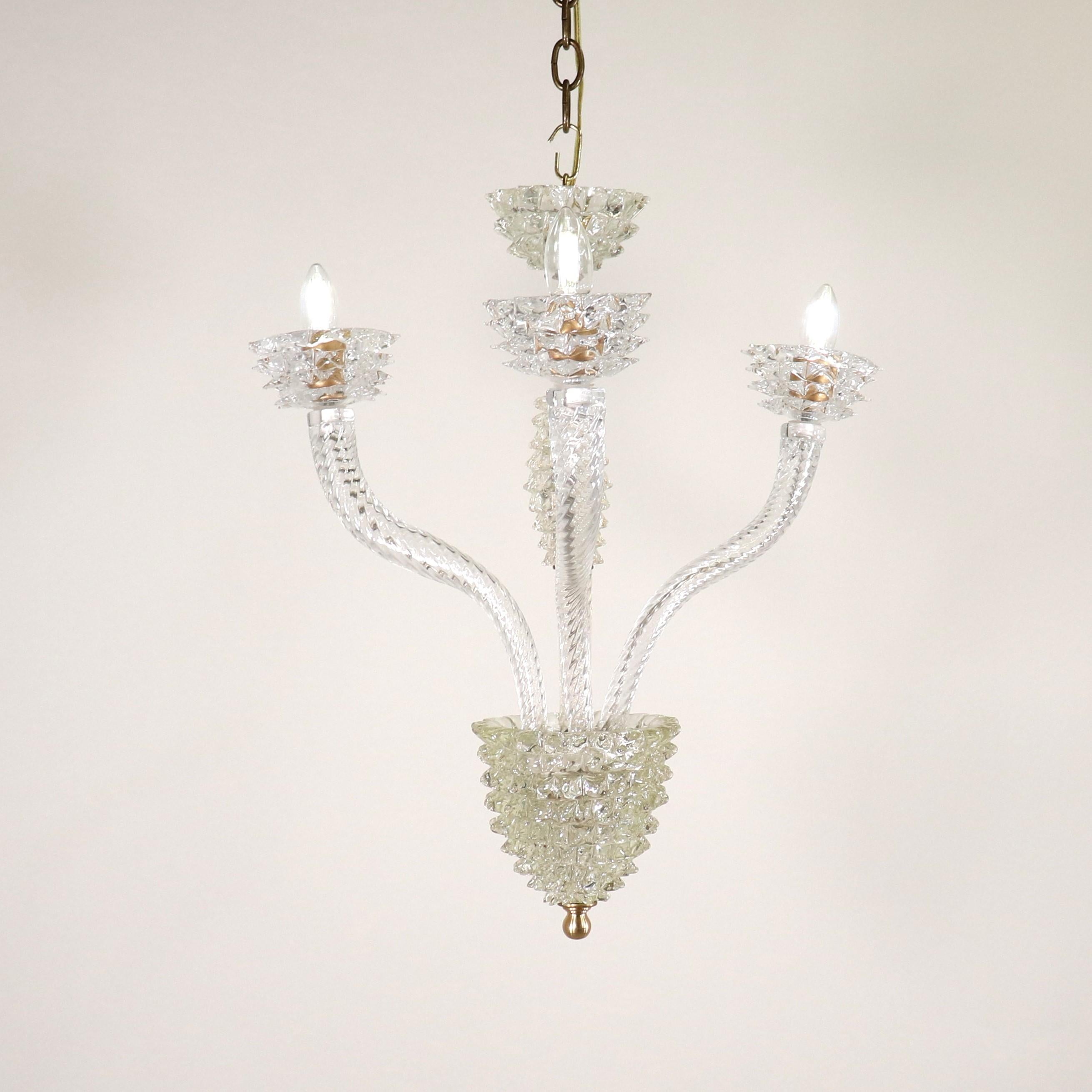This small but striking mid-century modern Murano chandelier has three scroll arms and features rostrato detailing in the manner of Ercole Barovier. The body and arms of this piece are made of clear, colorless Venetian glass. The various techniques