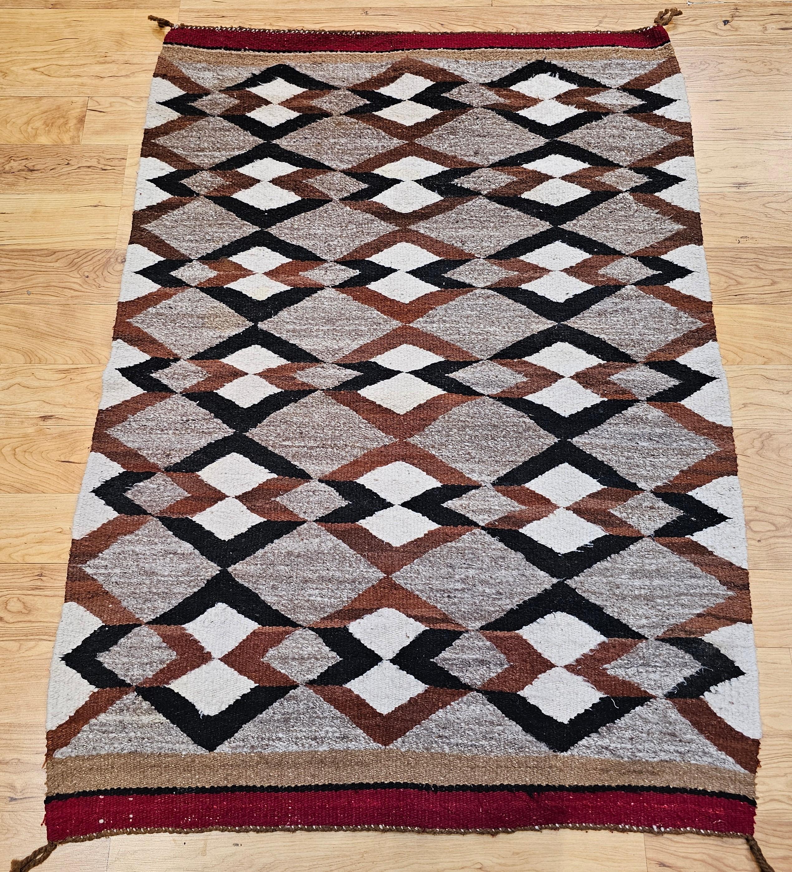 This vintage Native American Navajo area rug, woven circa the mid-1900s, features a bold and traditional diamond pattern in a warm, earthy palette of gray, ivory, brown, and black. A striking example of Navajo textile artistry, this rug embodies the