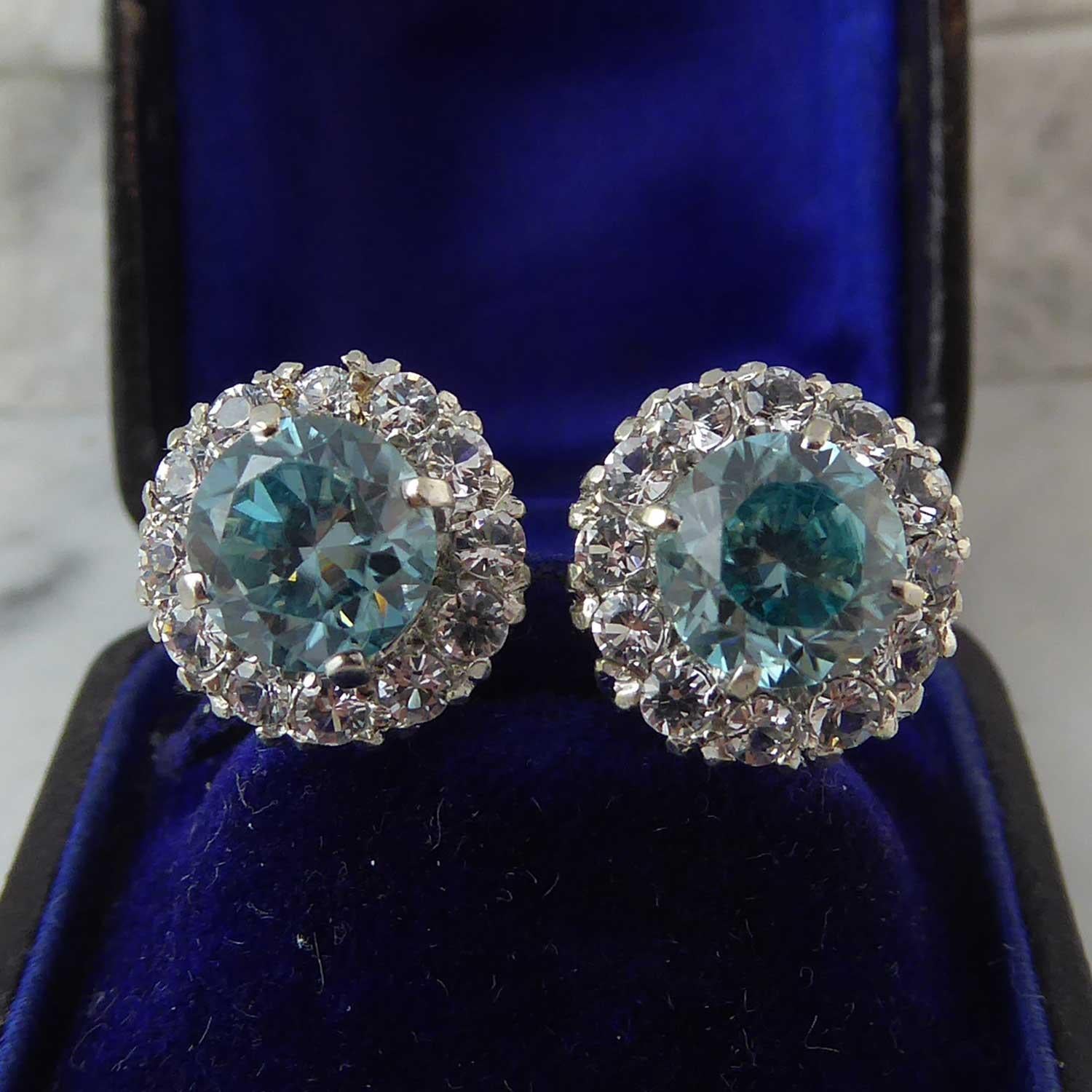 A pair of vintage natural blue zircon earrings in a surround of white stones assessed as white sapphire.  Centrally set with a faceted round blue zircon in four white claw setting to a surround of 11 white stones to an open basket cluster mount. 