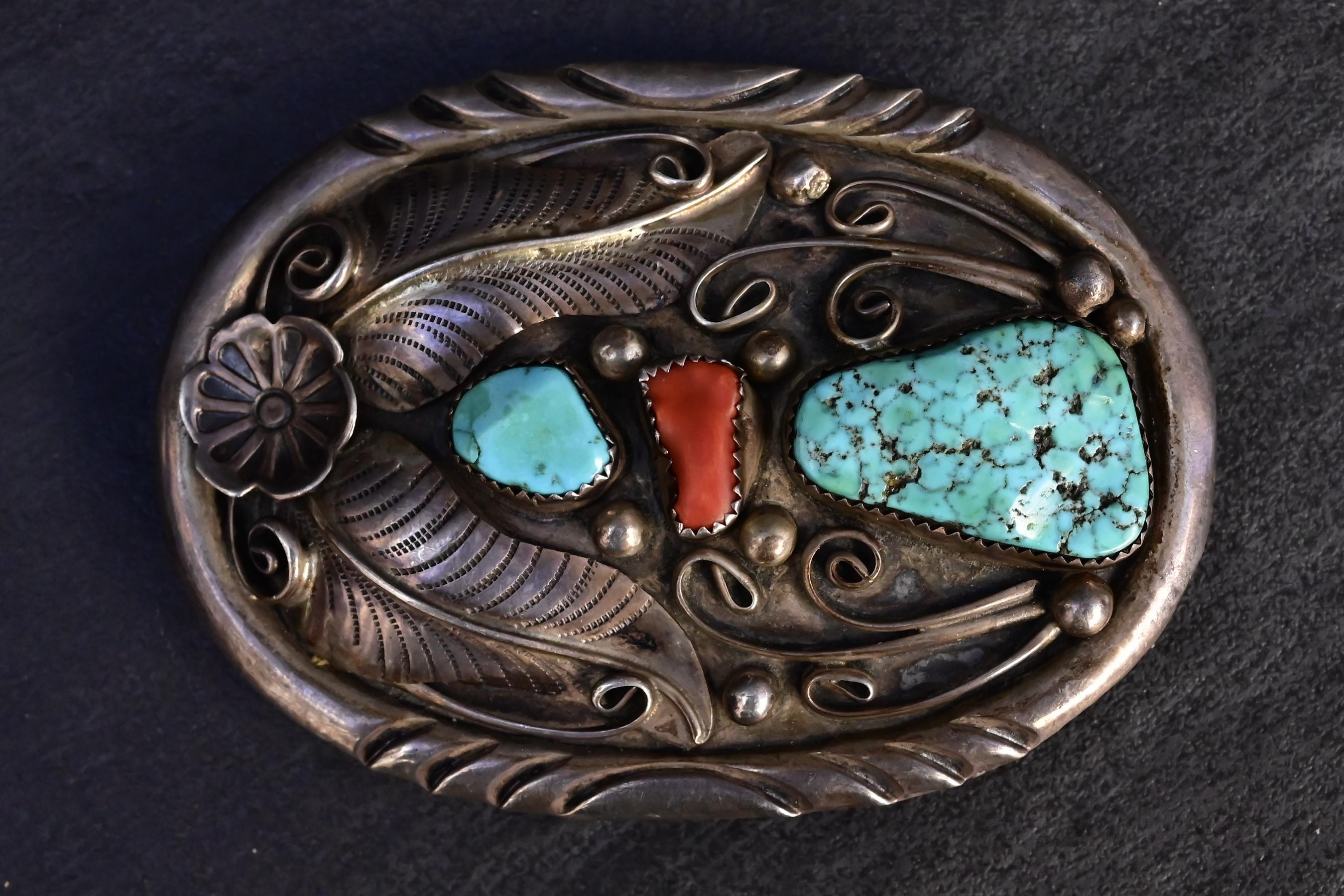 Diese auffällige Navajo-Gürtelschnalle ist aus massivem Sterlingsilber gefertigt und zeigt klassische handgeprägte Motive, wirbelnde Applikationen und traditionelle Blumenmotive. Die Schnalle ist mit zwei natürlichen Türkissteinen besetzt - einem