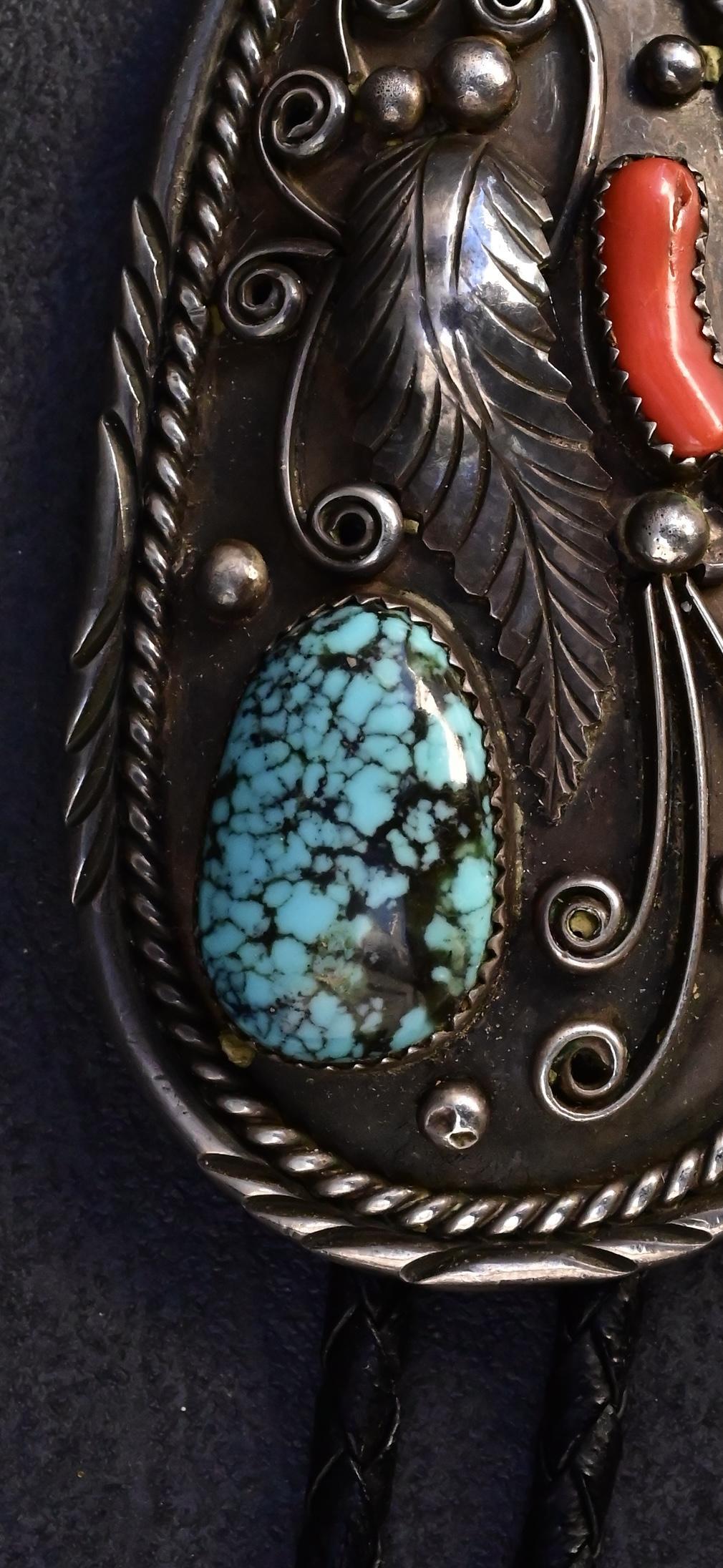 This striking vintage Navajo bolo tie features a beautifully handcrafted sterling silver slide adorned with natural turquoise and red coral cabochons. Traditional Navajo silverwork—including hand-stamped feather motifs, rope details, and flowing