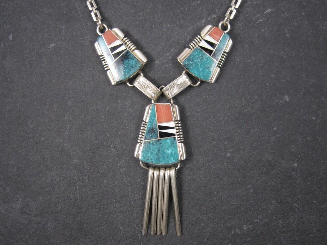 This gorgeous sterling silver necklace is the creation of Navajo silversmith John Charley.

It features inlay in pink spiny oyster, turquoise, jet and white shell.

This necklace measures 16 inches from end to end and 10 inches when clasped from