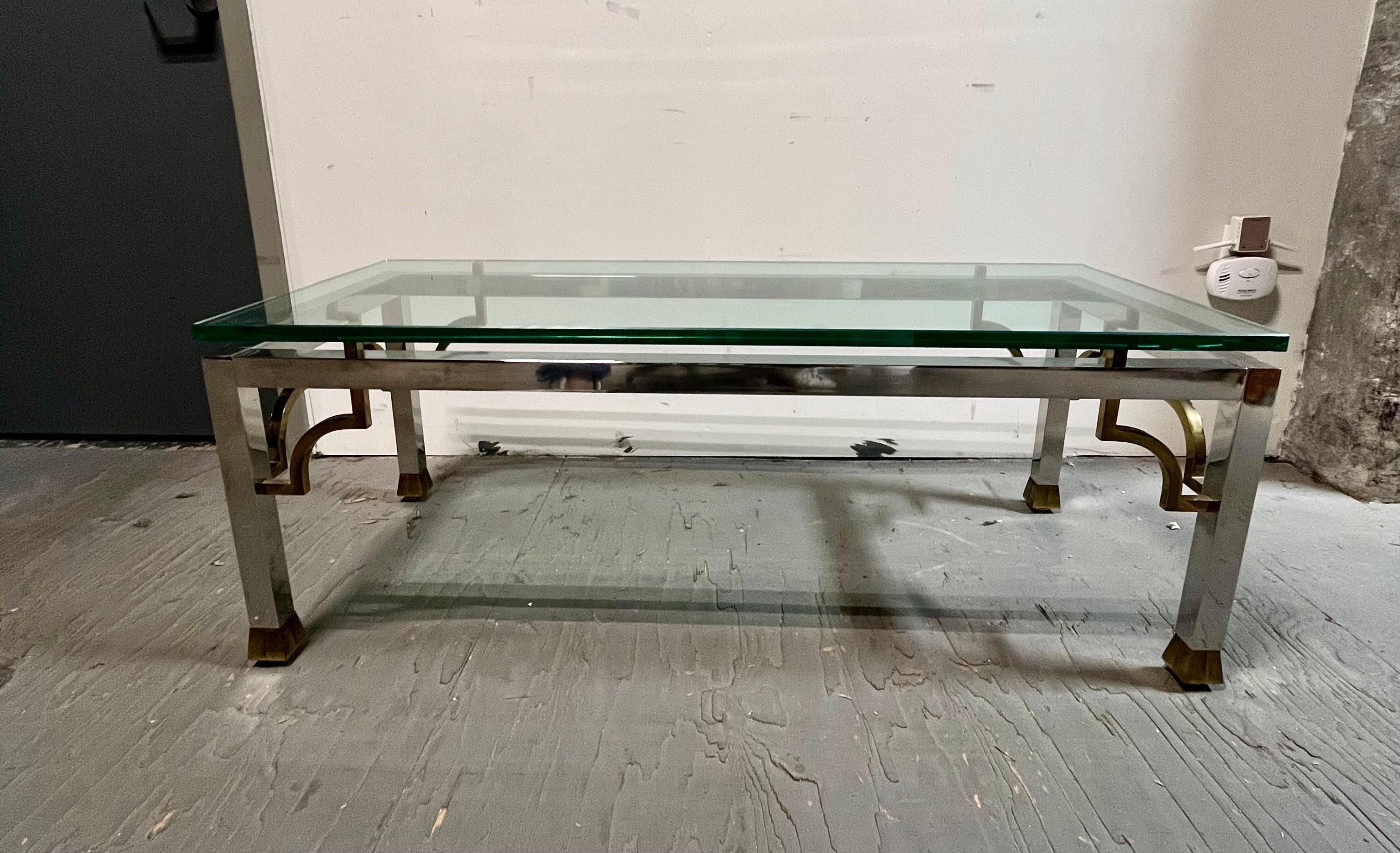 Magnifique table basse moderne de style néoclassique. Chrome avec des accents en laiton pour inclure des éléments de clé grecque. Les pieds joliment évasés ajoutent une touche unique. Le verre épais et lourd repose sur des cubes en laiton