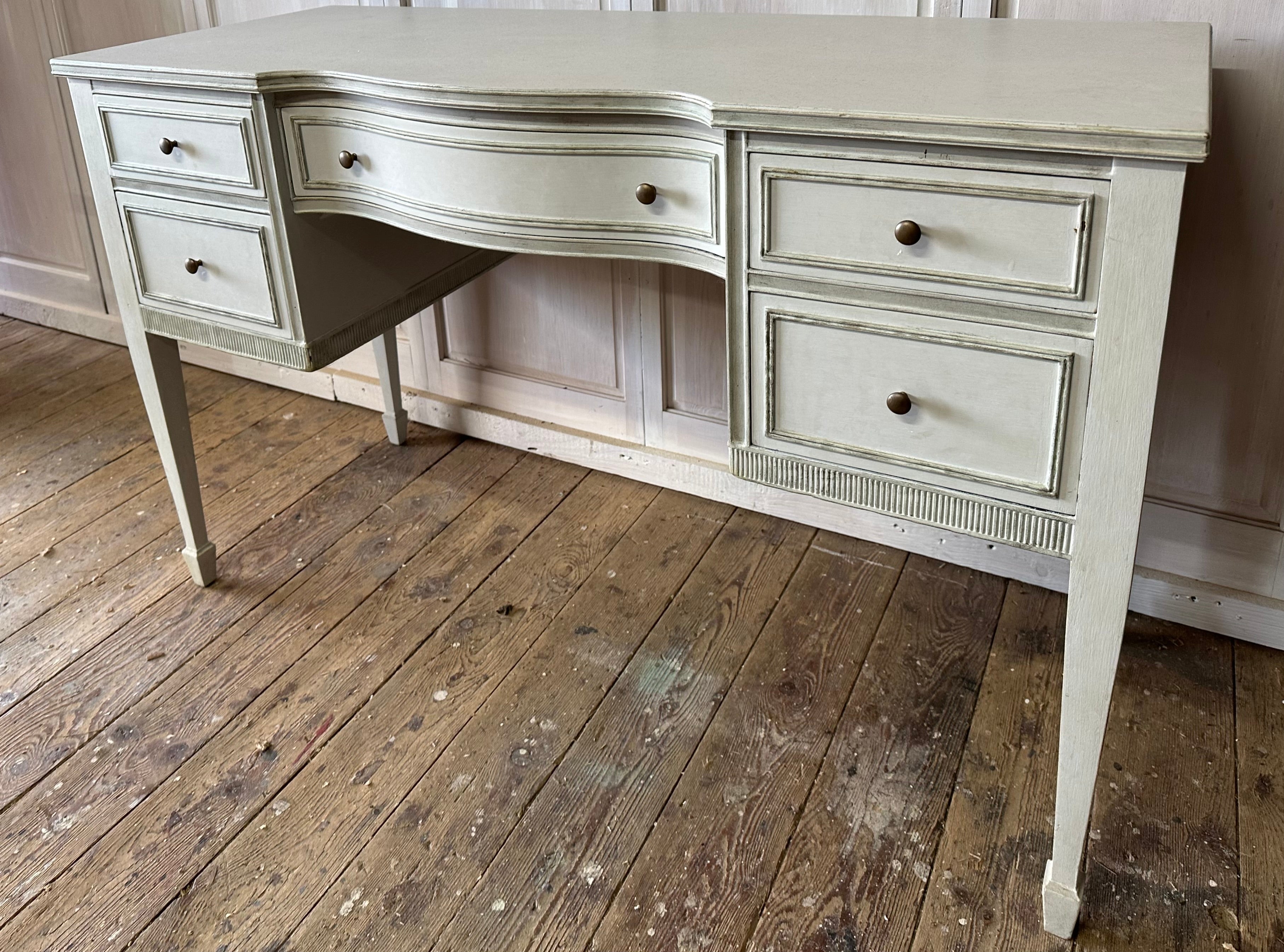 The vintage neoclassical painted dressing table, vanity, desk or night table has gilt detailing, serpentine front, brass pulls and tapered legs. Drawers are dovetailed. Can also be used as side table or nightstand. Painted a very light grey.  Label