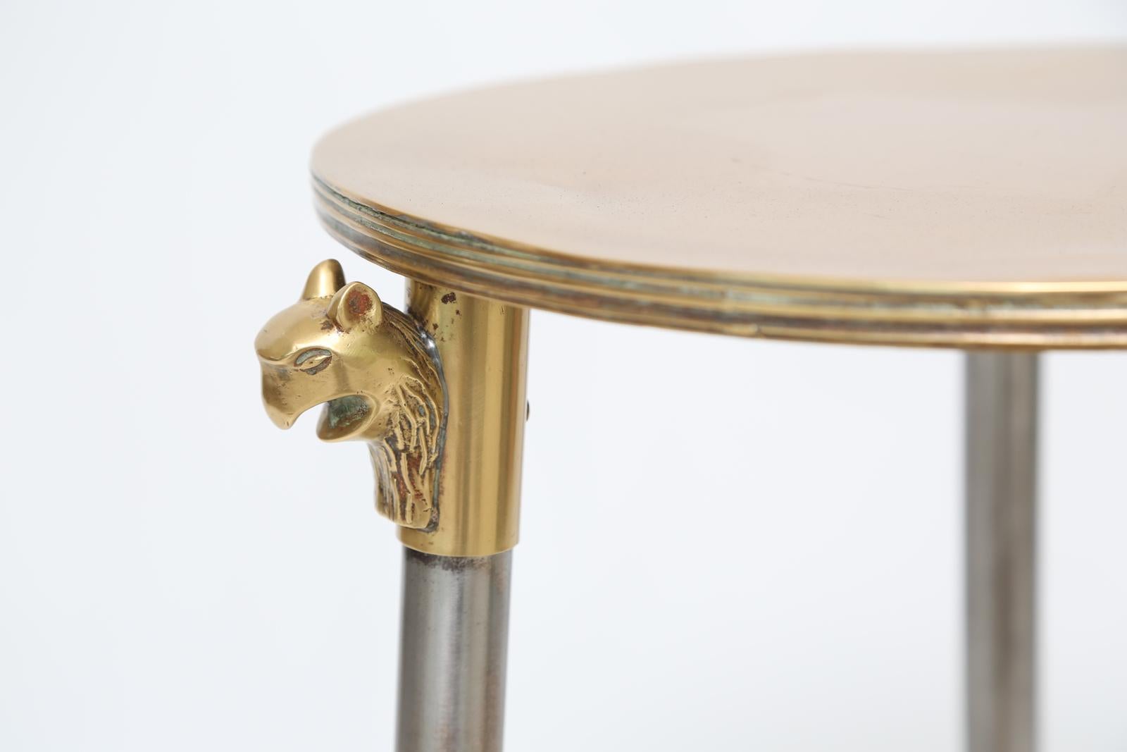 Candlestand side table, of polished brass and steel, having a round top, raised on a trio of panther-headed, round legs, joined by a shelf stretcher with concave sides, ending in feet fashioned as tassels. 

The circular top measures 13.25