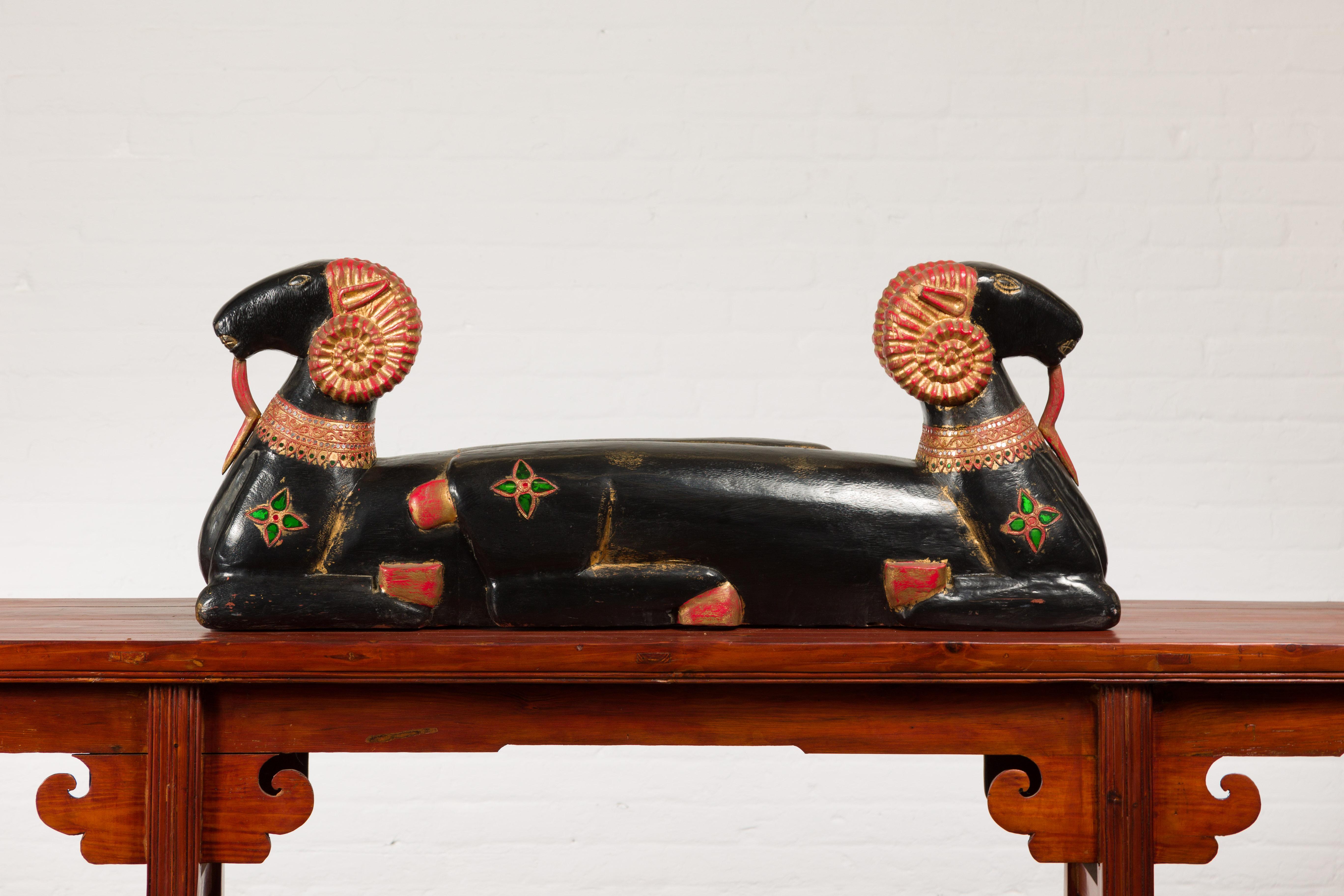 A vintage Northern Thai double ram black painted sculpture from the mid-20th century, with elegant antlers, green glass jewelry motifs and red accents. Found in Northern Thailand, this horizontal sculpture from the Midcentury period features two