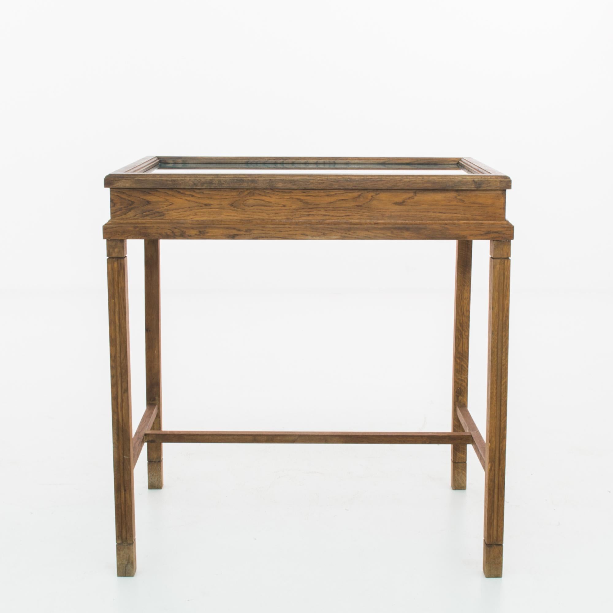 A display table from 1950s Germany. An oak display case with a glass top sits atop four slender legs, joined by an H-shaped strut. The wood has a rich brunette tone and a gentle shine; a symmetrical grain provides an organic decorative touch.