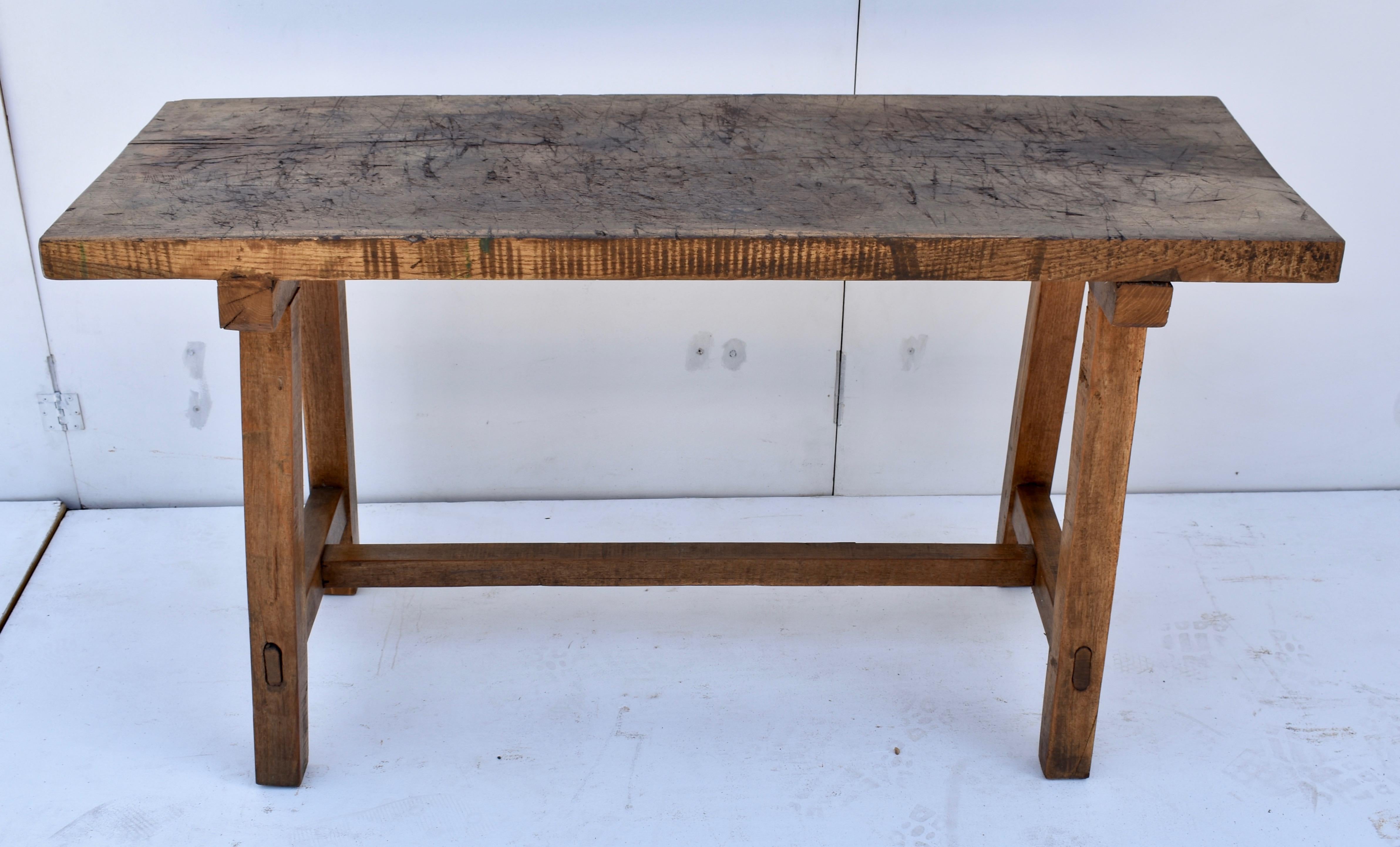 The top of this unusual Pig bench is a single slab of oak over two inches thick, gnarled and chopped through decades of purposeful use. The base consists of two trestles, mortised into a cross member at the top and joined at the bottom with oak